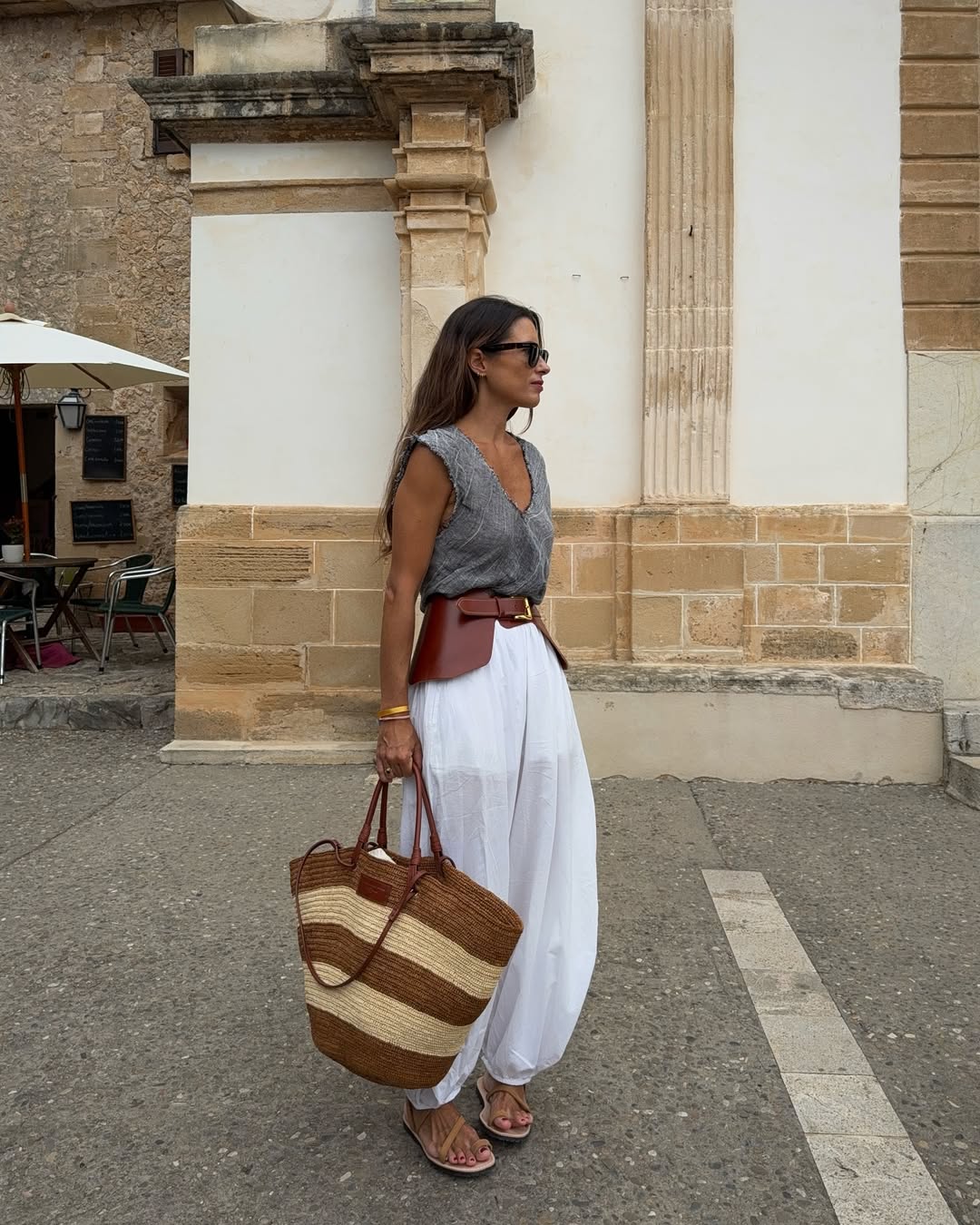 Foto de uma mulher morena usando um look composto por regata cinza, cal&ccedil;a de linho branca, cinto de couro marrom e bolsa de palha listrada