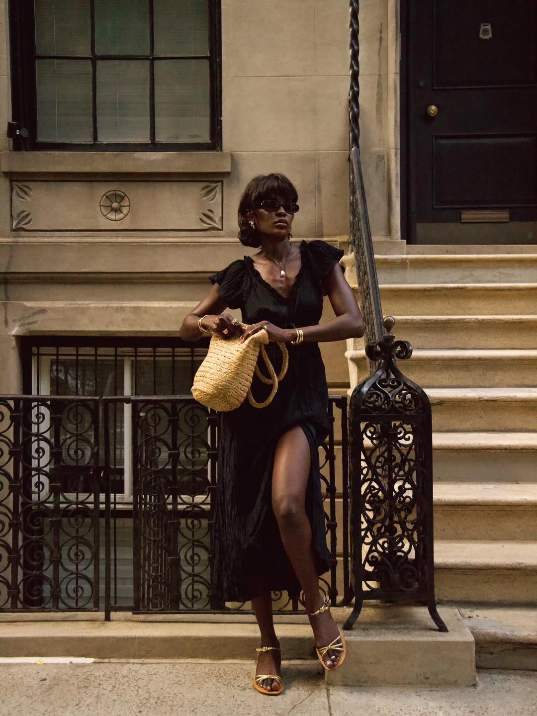 Foto de uma mulher negra usando um look composto por vestido longo preto, bolsa de palha e &oacute;culos de sol