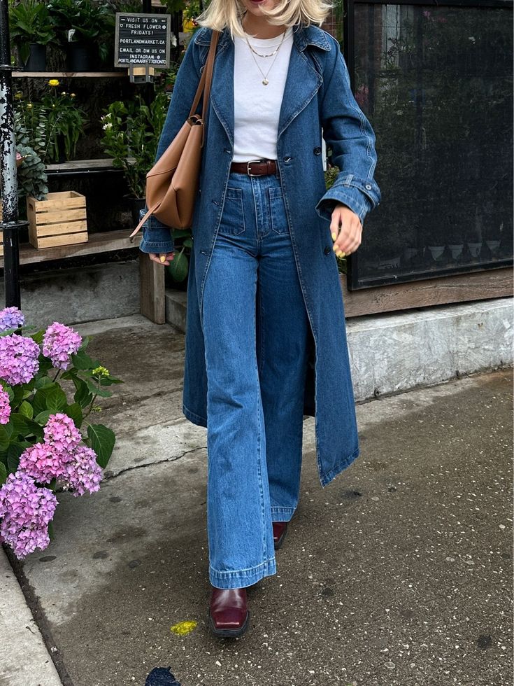 Foto de um look composto por regata branca, jaqueta jeans longa, cal&ccedil;a jeans, bolsa marrom, cinto marrom e bota de couro