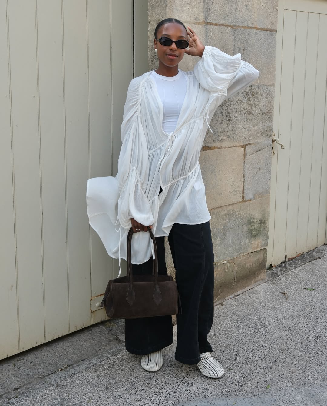 Foto de uma mulher negra usando um look composto por blusa branca longa, cal&ccedil;a jeans preta, bolsa marrom e t&ecirc;nis branco