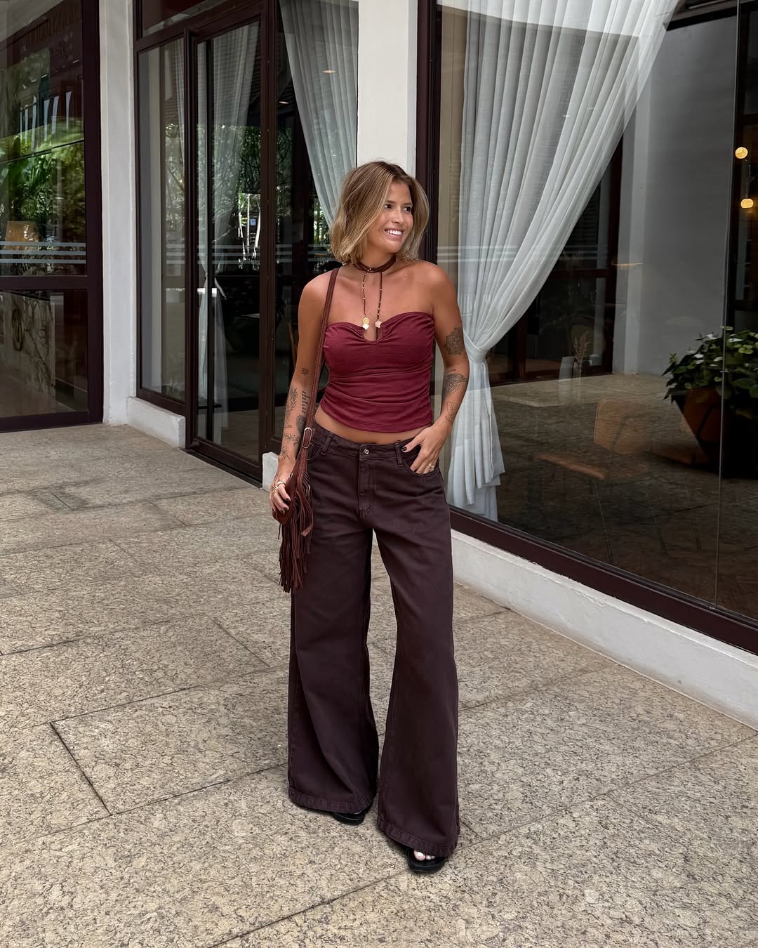 Foto de uma mulher loira usando um look composto por blusa vinho, cal&ccedil;a marrom e bolsa