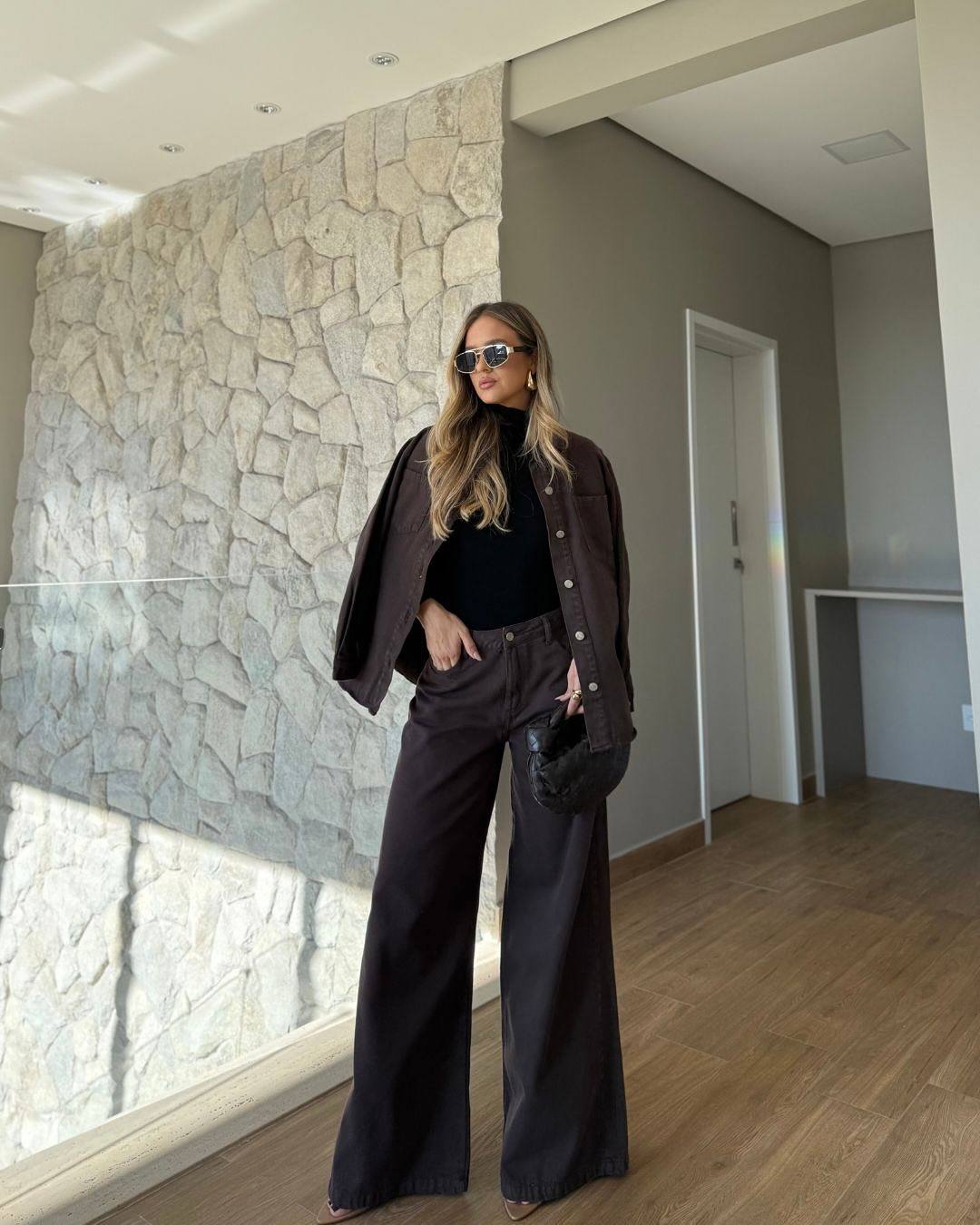 Foto de uma mulher loira usando um look composto por blusa de gola alta preta, jaqueta marrom, &oacute;culos de sol, cal&ccedil;a marrom e bolsa