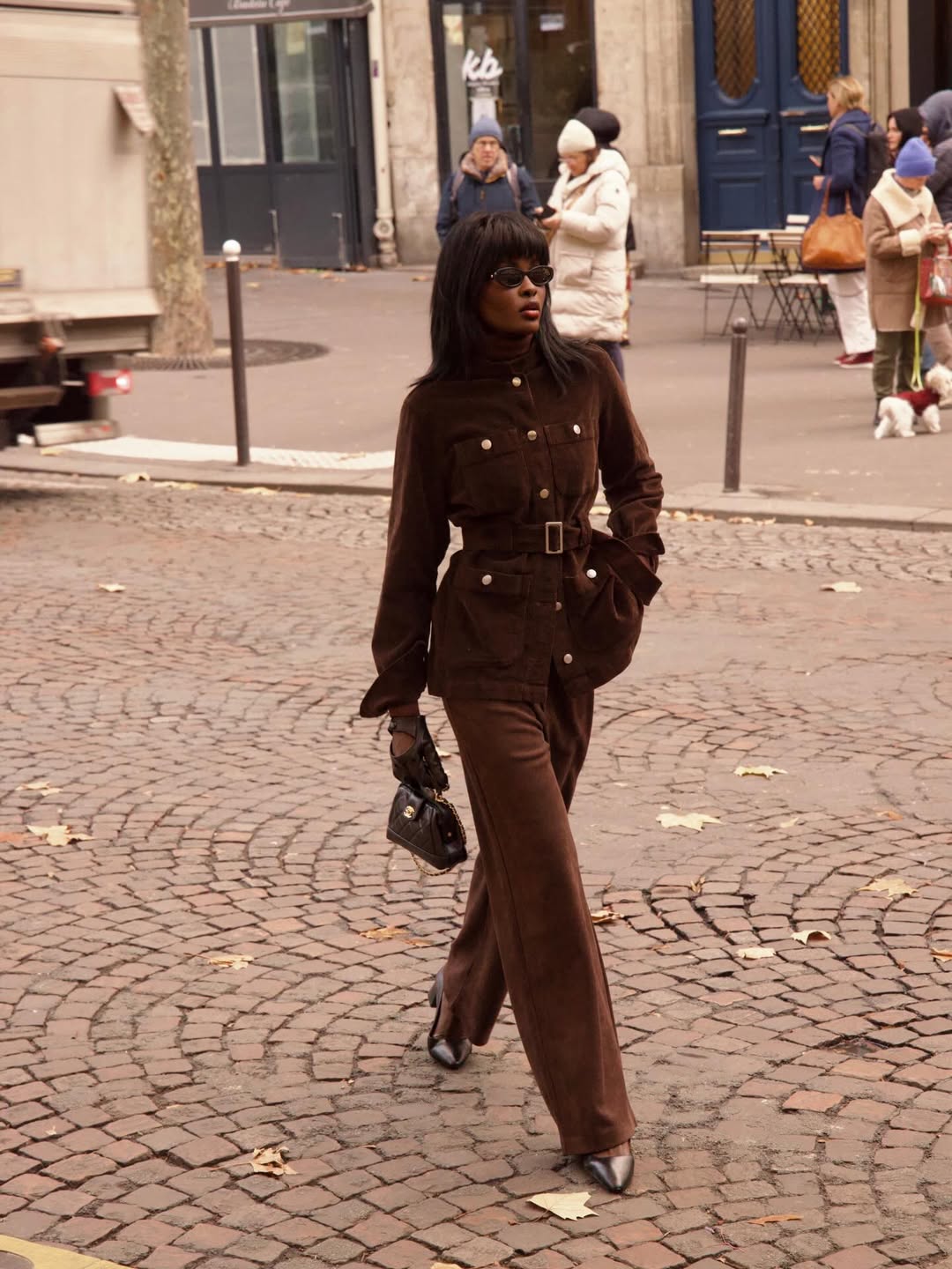 Foto de uma mulher negra usando um look composto por jaqueta, cal&ccedil;a, bolsa e salto, todos marrom