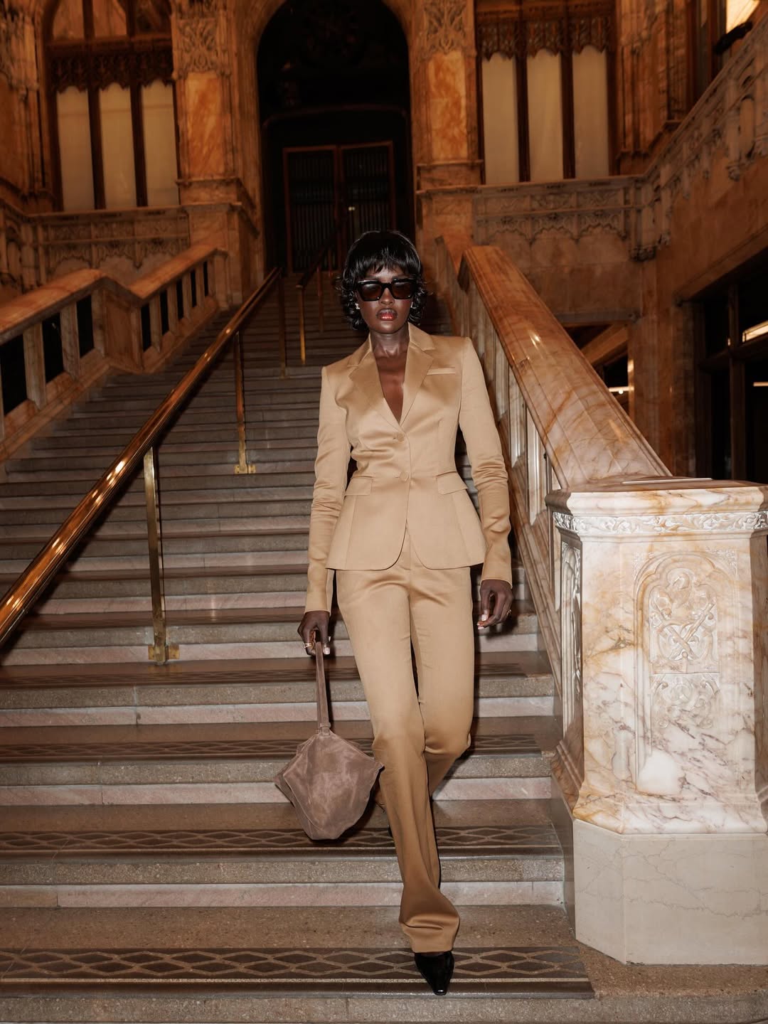 Foto de uma mulher negra usando um look composto por blazer e cal&ccedil;a de alfaiataria em tom areia brilhante, &oacute;culos de sol, bolsa e salto