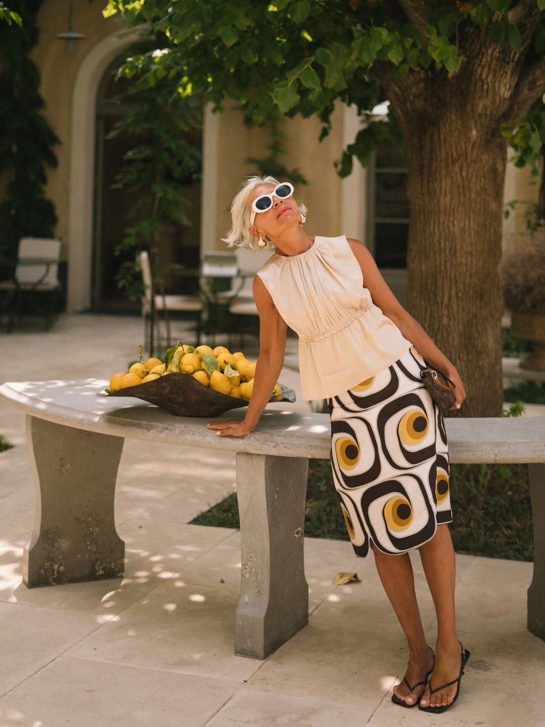 Foto de uma mulher branca, com mais de 60 anos, usando um look composto por regata de alfaiataria off white, saia branca, amarela e preta, &oacute;culos de sol e sand&aacute;lia havaianas