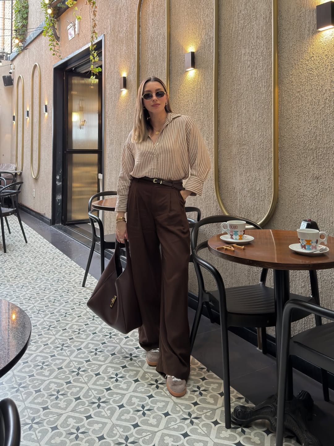 Foto de uma mulher loira usando um look composto por camisa, cal&ccedil;a marrom, bolsa marrom e t&ecirc;nis