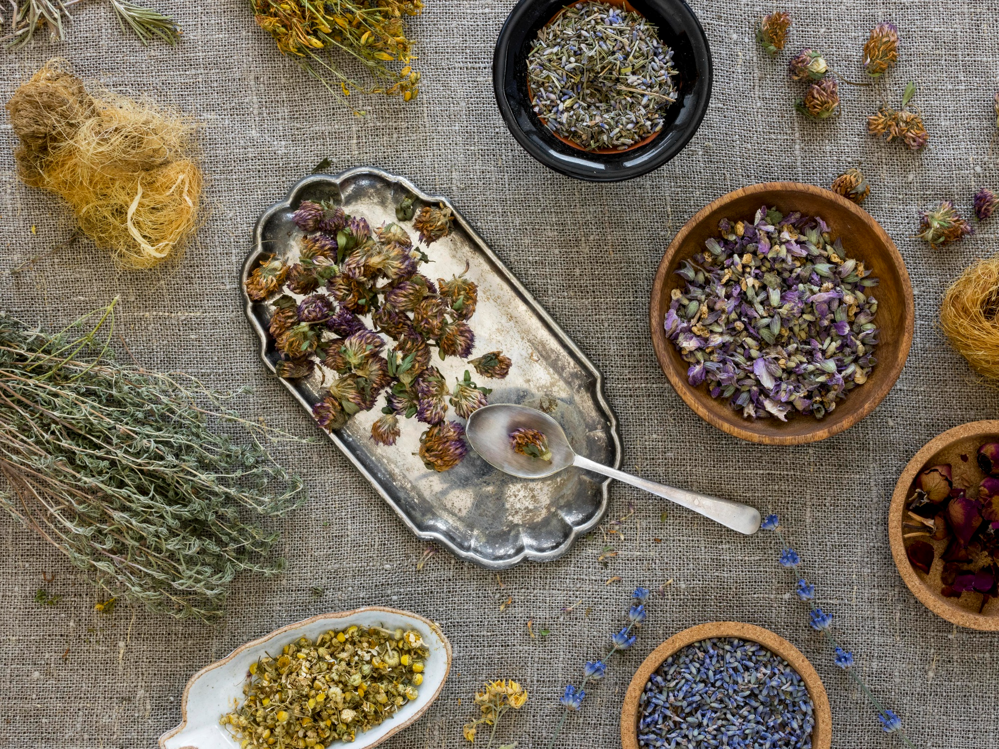 Ervas secas como lavanda, camomila, alecrim e flores variadas organizadas em tigelas e bandeja sobre tecido r&uacute;stico, representando ingredientes para banho de limpeza energ&eacute;tica e renova&ccedil;&atilde;o p&oacute;s-Carnaval.
