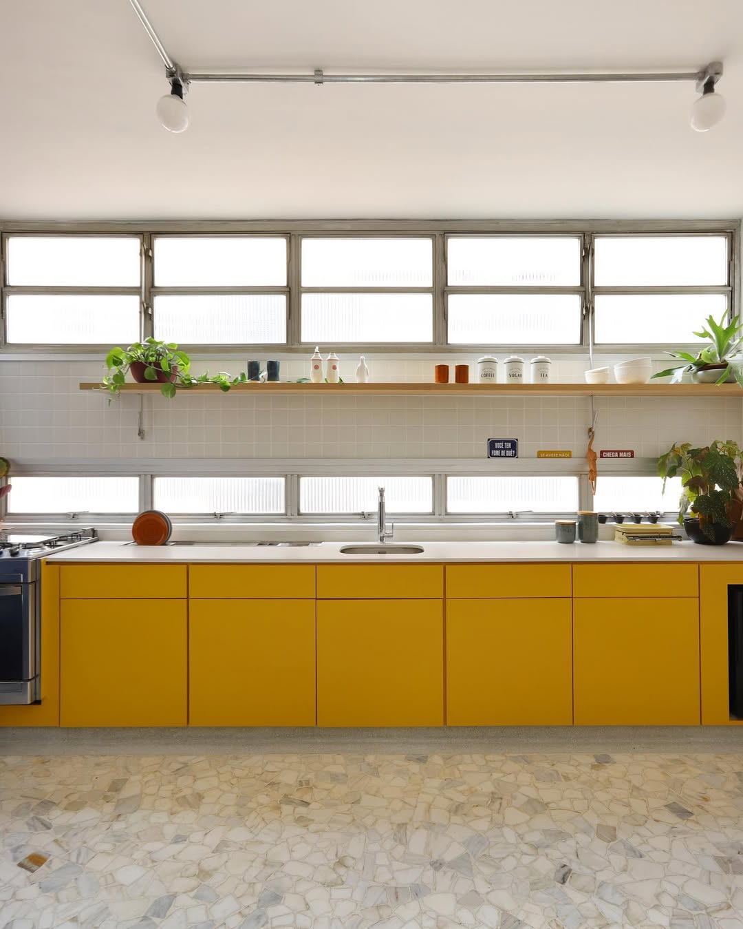 Cozinha com bancada comprida, arm&aacute;rios amarelos, janelas atr&aacute;s da bancada e piso de caquinho