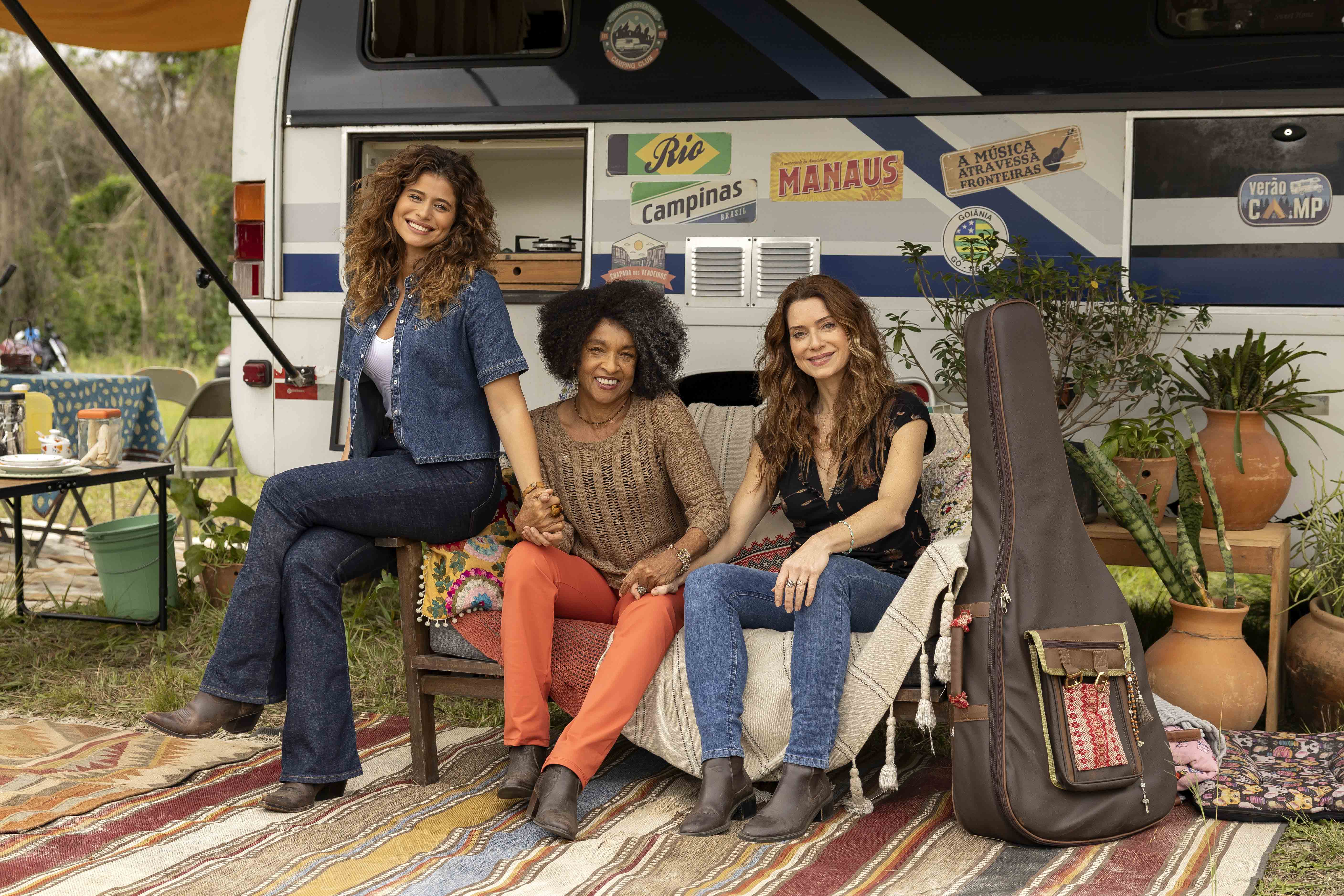 Agrado posa sorridente ao lado de duas mulheres em frente a um trailer de viagem, com violão ao lado, em clima de estrada e bastidores da turnê em Coração Acelerado