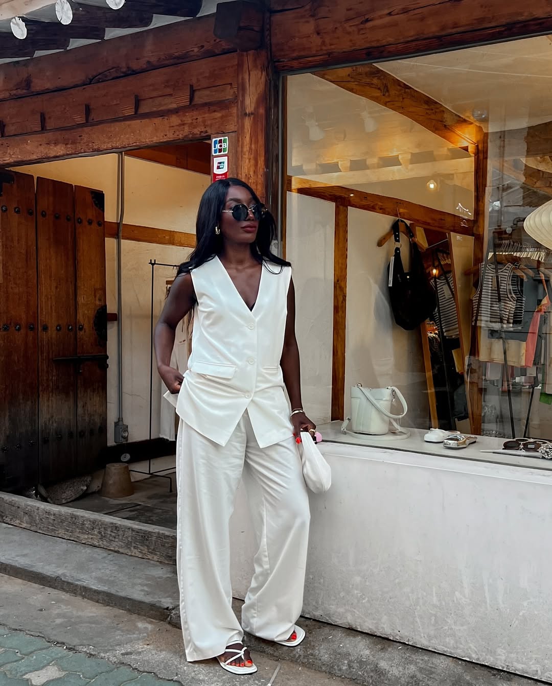 Foto de uma mulher negra usando um look composto por colete de alfaiataria branco, cal&ccedil;a branca, bolsa e sand&aacute;lia