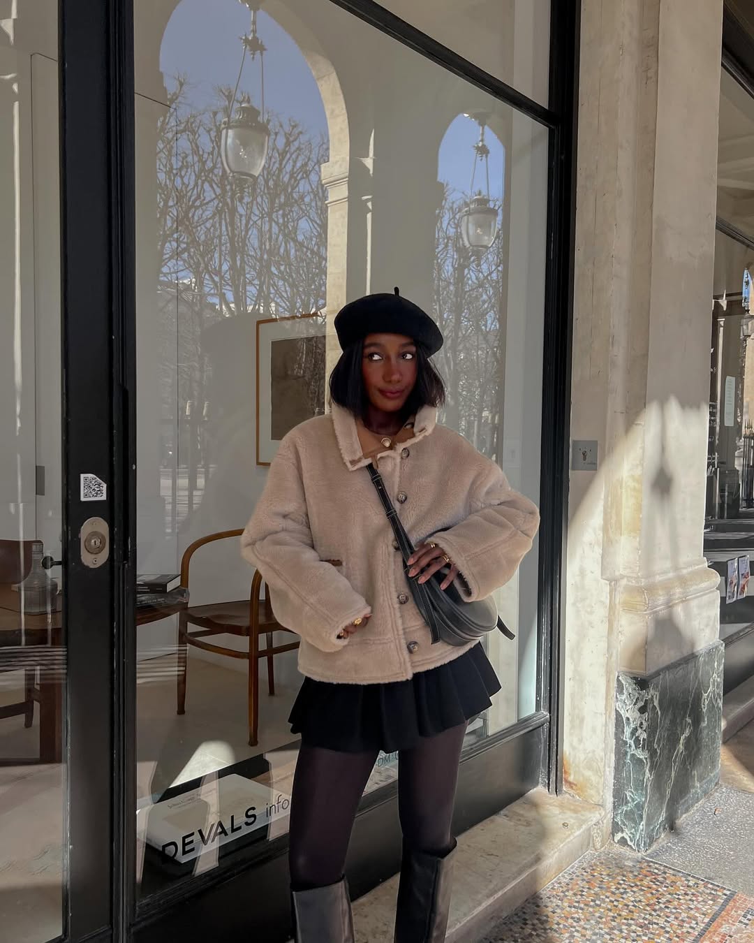 Foto de uma mulher negra usando um look composto por blusa de frio marorm, saia plissada preta, bota de couro preta, bolsa e boina