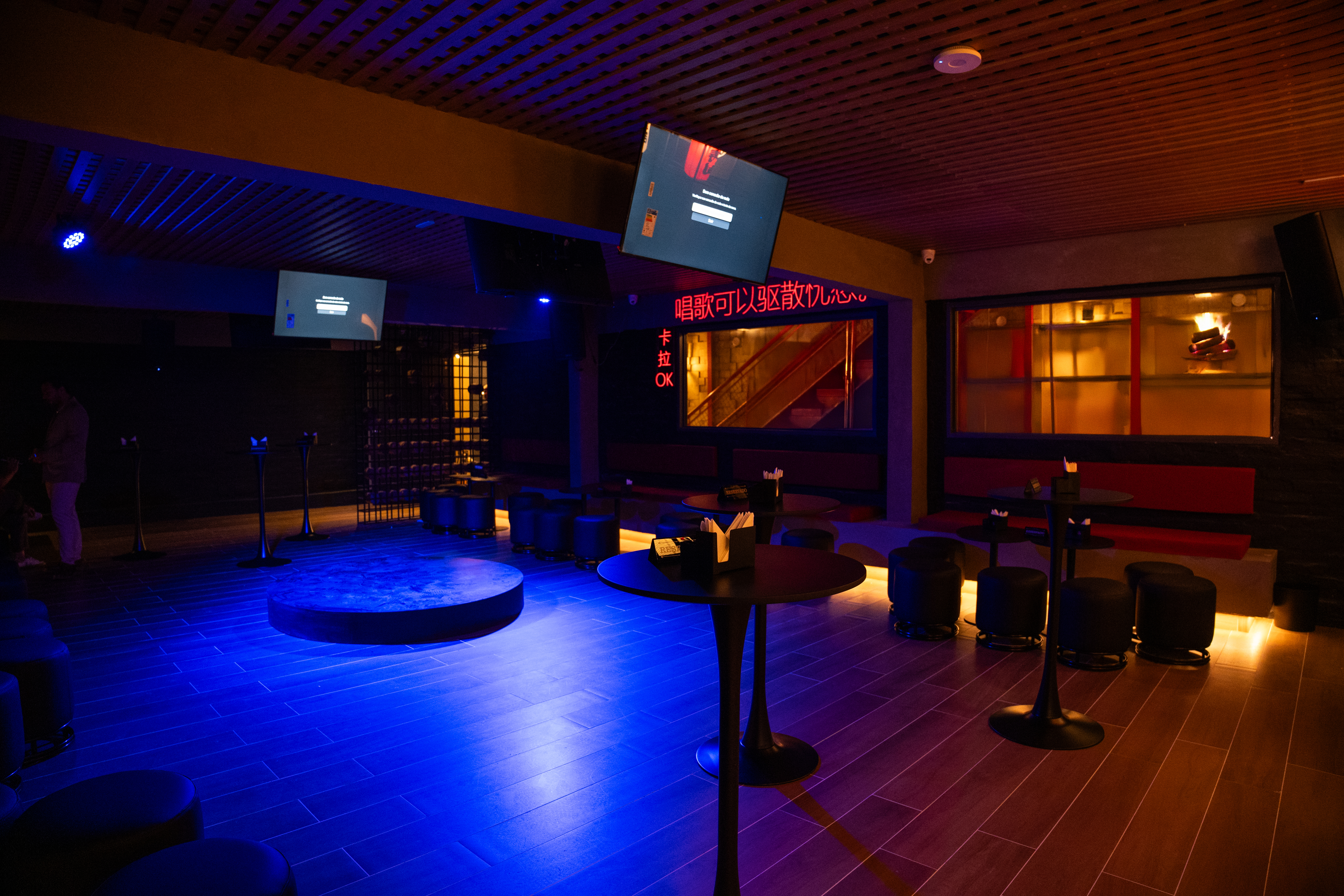 Lounge da nova unidade do Yellow K em Pinheiros, S&atilde;o Paulo, com mesas altas, ilumina&ccedil;&atilde;o em neon azul e vermelho, telas para karaok&ecirc; e ambiente inspirado em bares asi&aacute;ticos.