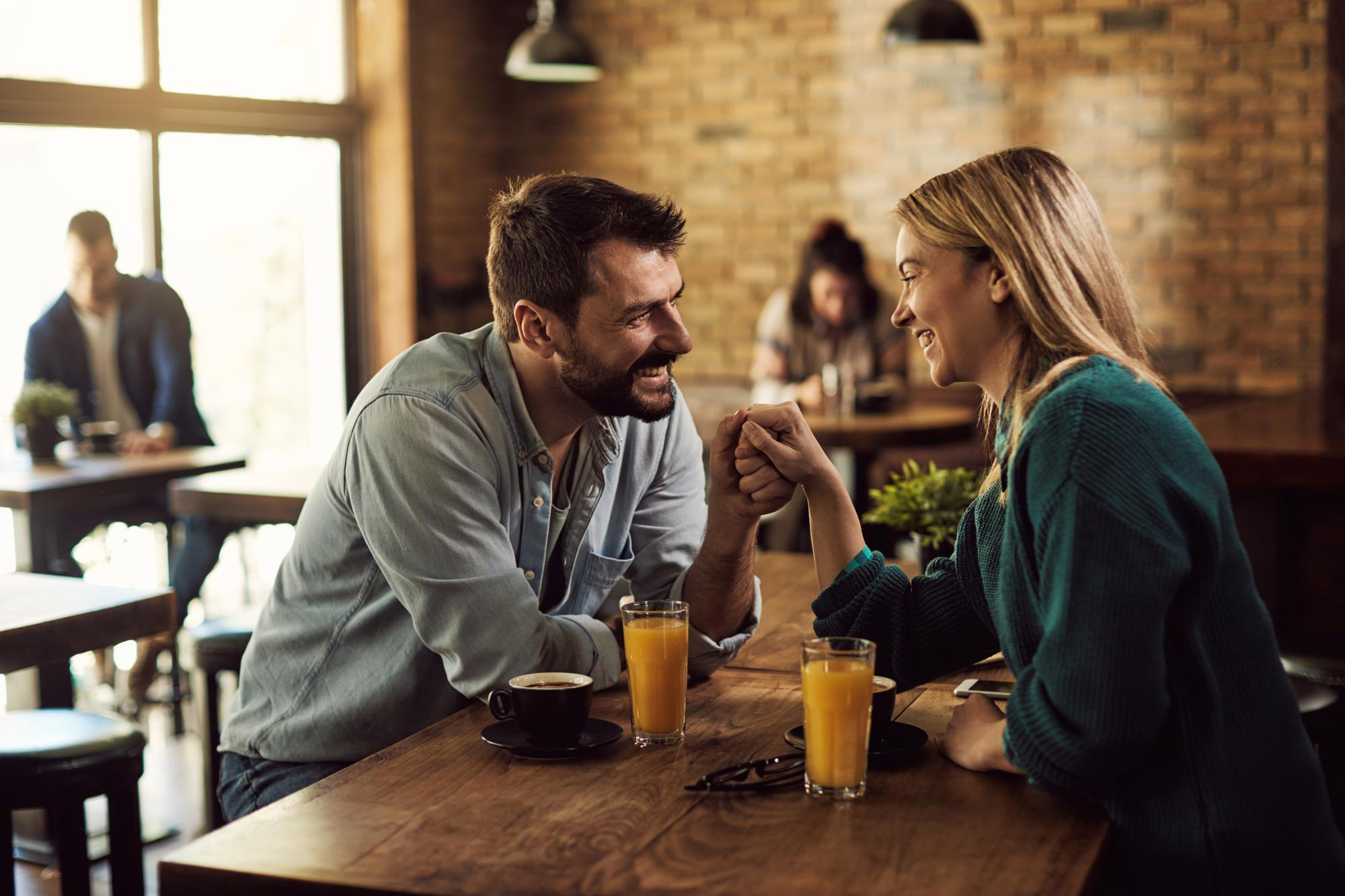 Casal sorrindo e segurando as m&atilde;os em um caf&eacute; iluminado, com clima &iacute;ntimo e descontra&iacute;do, representando conex&atilde;o, cumplicidade e as mem&oacute;rias afetivas de um relacionamento intenso.