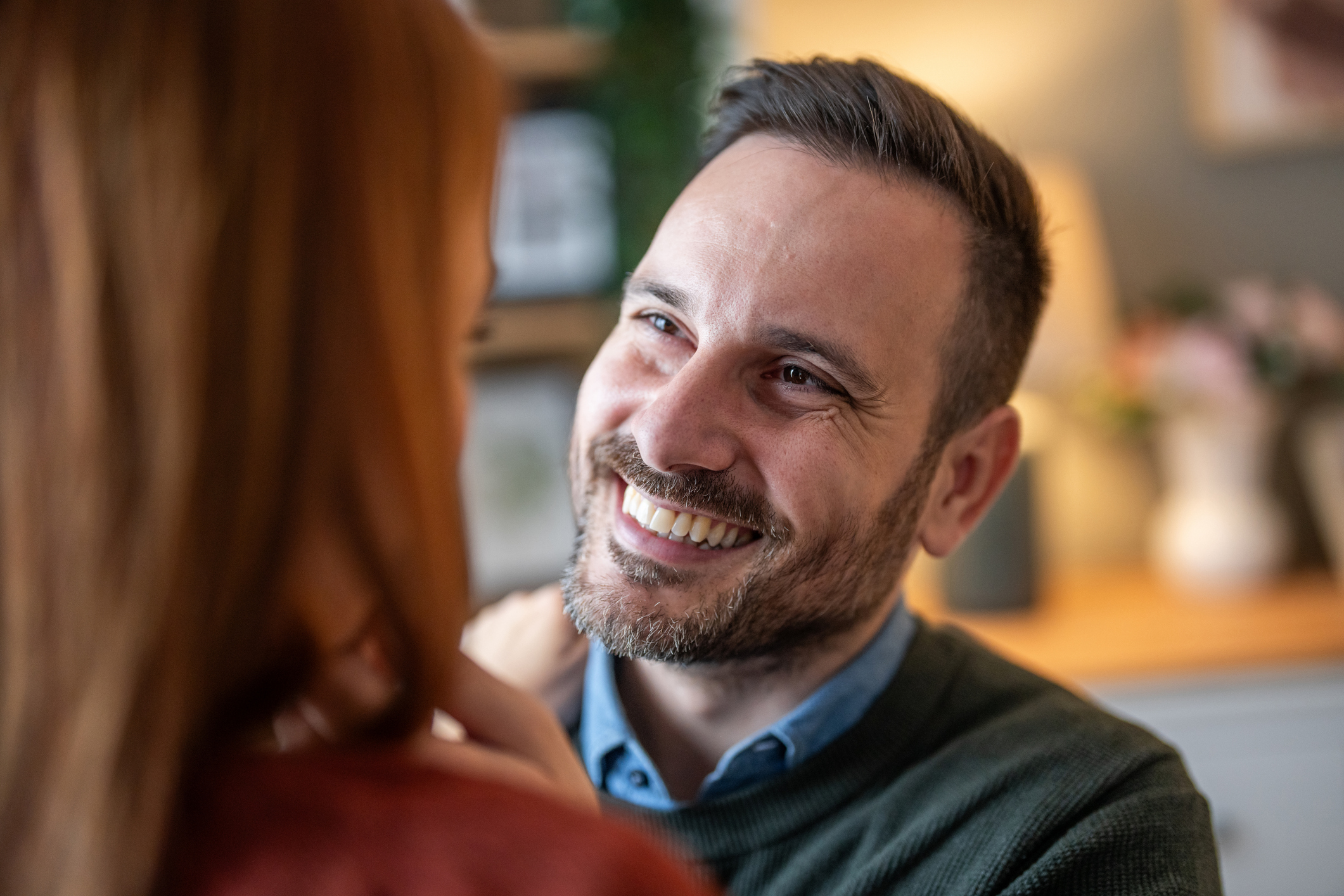 Homem sorrindo enquanto olha para uma mulher de perto, em um momento íntimo e afetuoso dentro de casa, com expressão de carinho e conexão emocional.