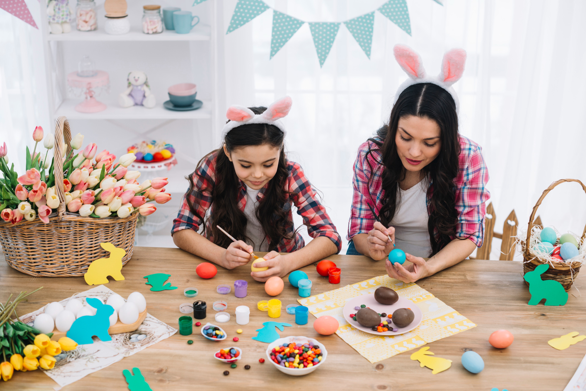 M&atilde;e e filha, ambas com orelhas de coelho, pintam ovos de P&aacute;scoa juntas em uma mesa cheia de materiais de artesanato e doces