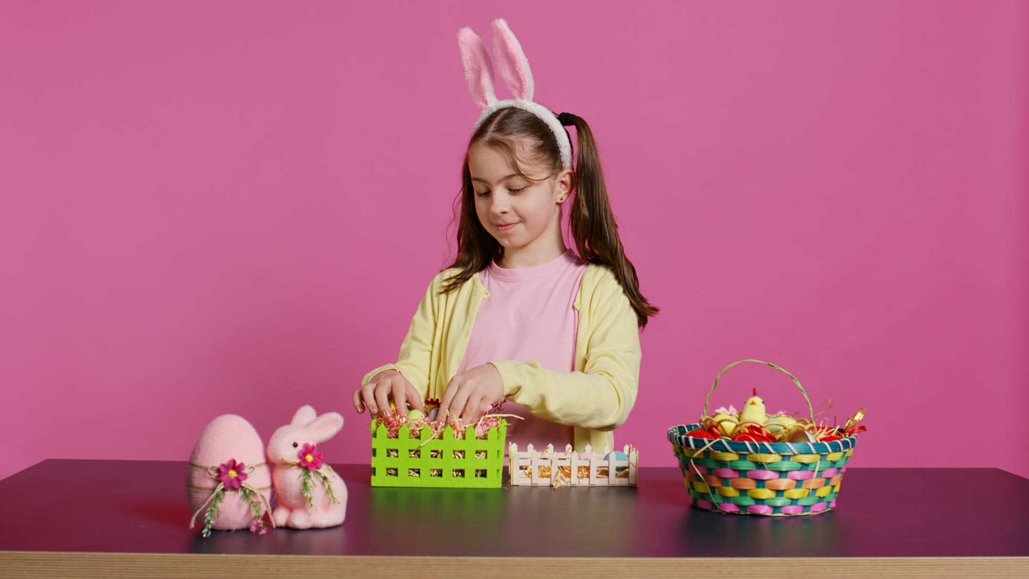 Menina usando tiara de coelho decora ovos de P&aacute;scoa em uma mesa com tintas, cestinhas e enfeites coloridos sobre fundo rosa.