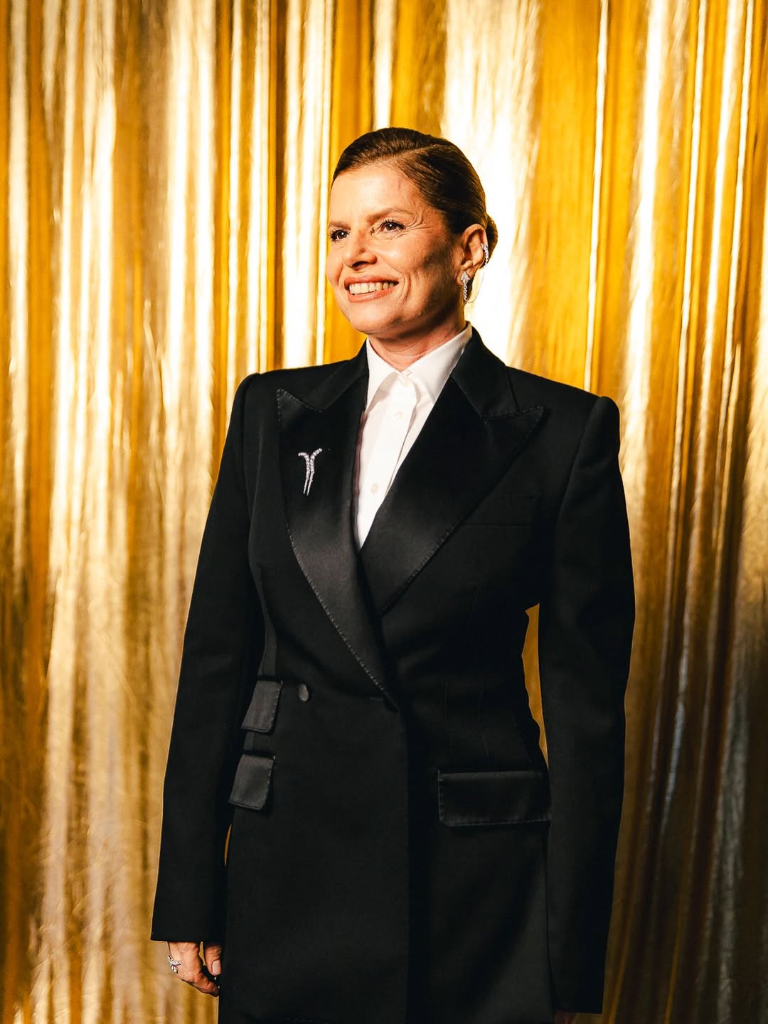 Foto de D&eacute;bora Bloch no Melhores do Ano. A imagem mostra seu tronco para cima. Ela usa o cabelo preso e veste um look composto por blazer preto, camisa branca e broche