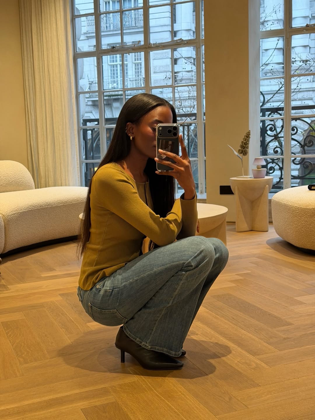 Foto de uma mulher negra, agachada, tirando foto em frente ao espelho. Ela usa uma blusa verde, cal&ccedil;a boot cut e salto