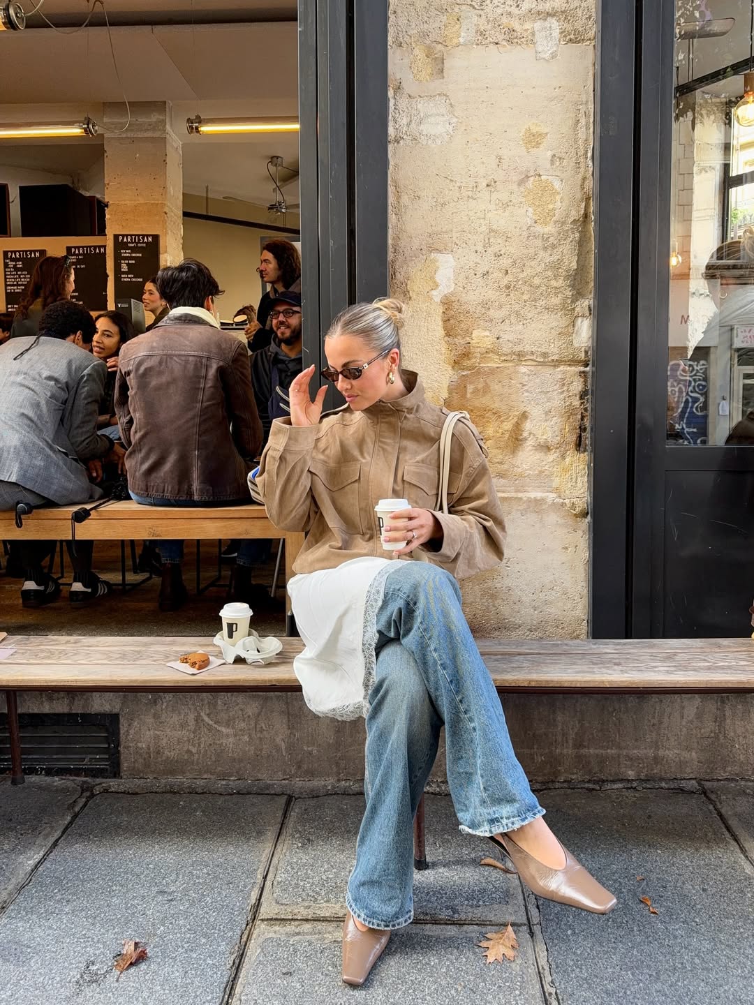 Foto de uma mulher loira usando um look composto por jaqueta marrom, cal&ccedil;a boot cut, len&ccedil;o no cabelo, &oacute;culos de sol e sapatilha prateada. Ela est&aacute; sentada em frente a um caf&eacute;