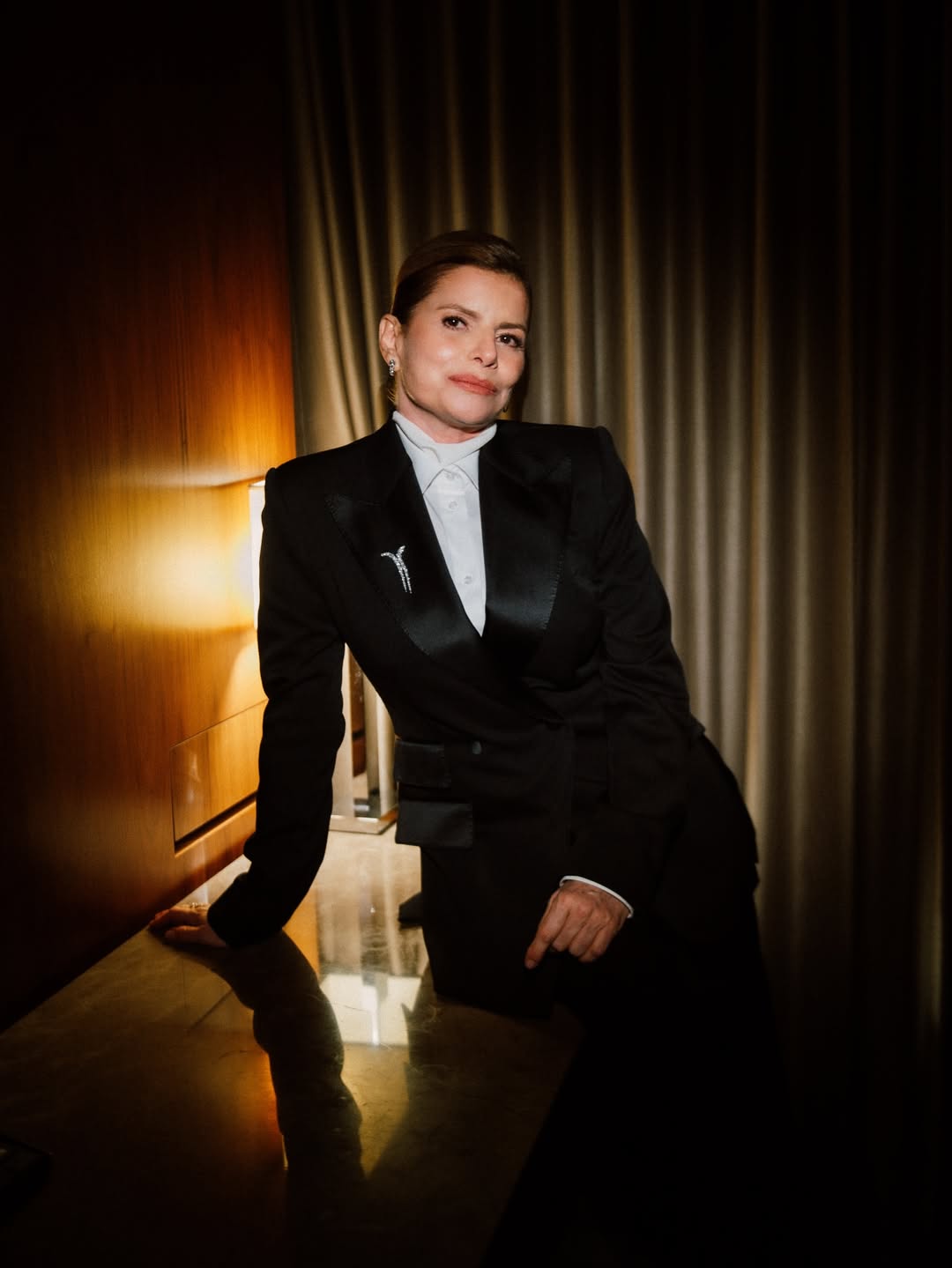 Foto de D&eacute;bora Bloch. Ela &eacute; branca, usa o cabelo preso e veste um look composto por blazer preto, cal&ccedil;a de alfaiataria preta, camisa branca e broche