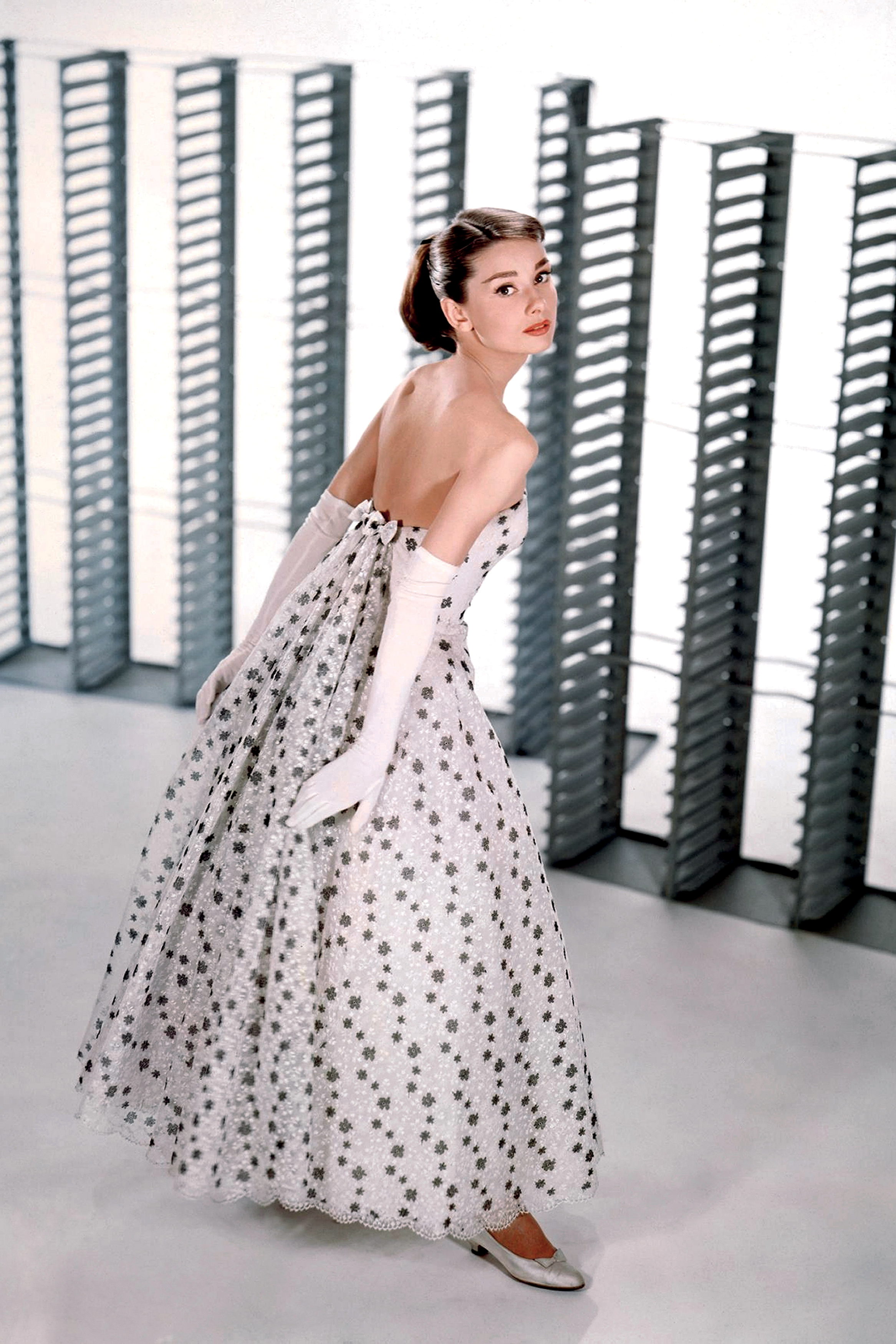 Audrey Hepburn posa com um elegante vestido de alta-costura, com saia ampla e luvas longas, refletindo a est&eacute;tica refinada que se tornou sua marca registrada e que marcou sua colabora&ccedil;&atilde;o com Givenchy no cinema.