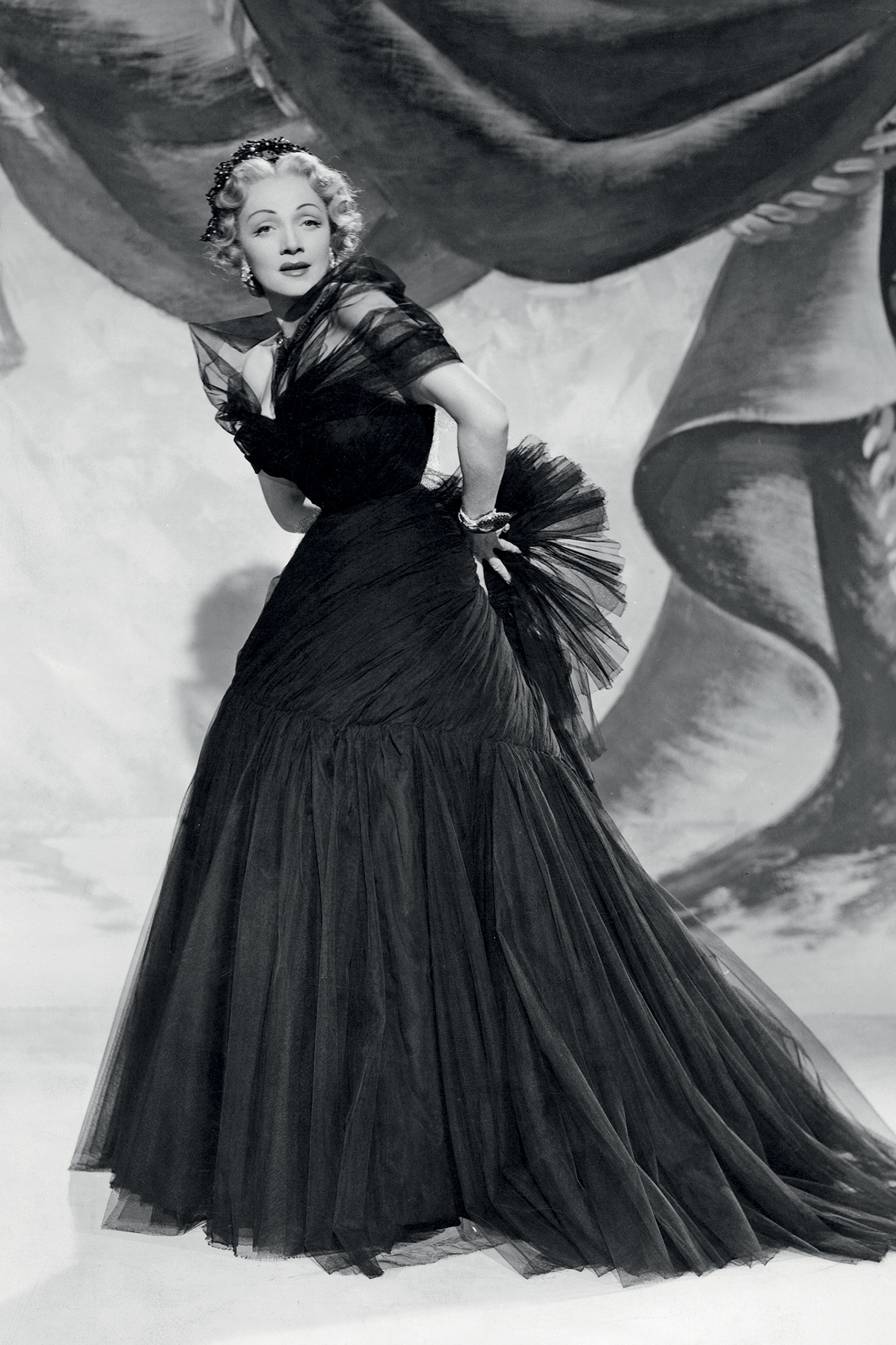 Marlene Dietrich aparece em um retrato cl&aacute;ssico usando um vestido de gala dram&aacute;tico e volumoso. O visual reflete a eleg&acirc;ncia intensa e teatral que caracterizava seu estilo e sua liga&ccedil;&atilde;o com a alta-costura.