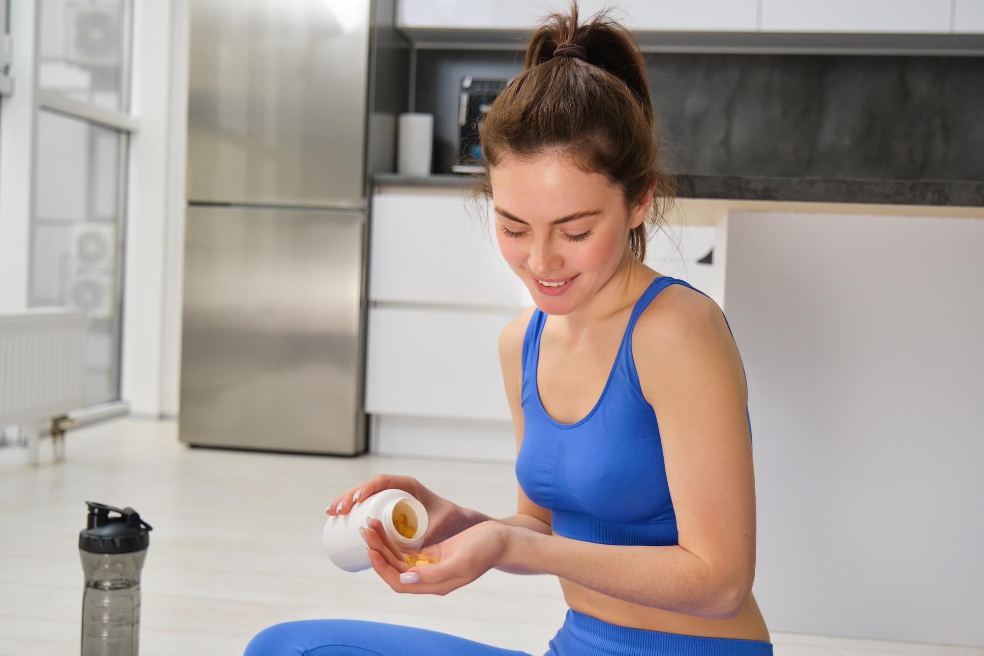 Mulher em roupa esportiva sentada em casa, segurando c&aacute;psulas de suplemento na m&atilde;o ao lado de uma garrafa de &aacute;gua, representando consumo de vitaminas na rotina fitness.
