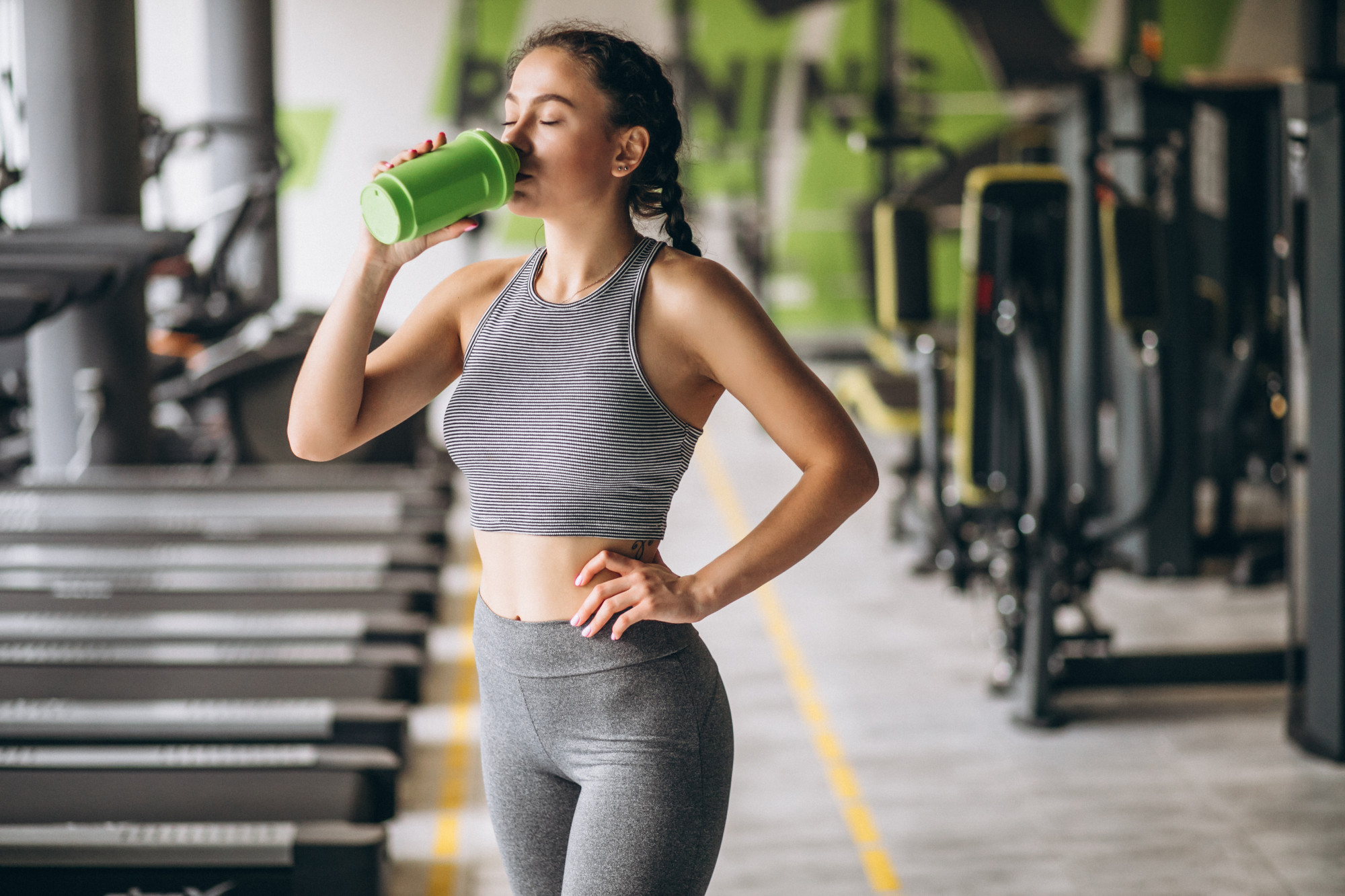 Mulher em academia vestindo roupa de treino, bebendo shake em garrafa verde ap&oacute;s exerc&iacute;cio, com equipamentos ao fundo, simbolizando uso de suplementa&ccedil;&atilde;o no p&oacute;s-treino