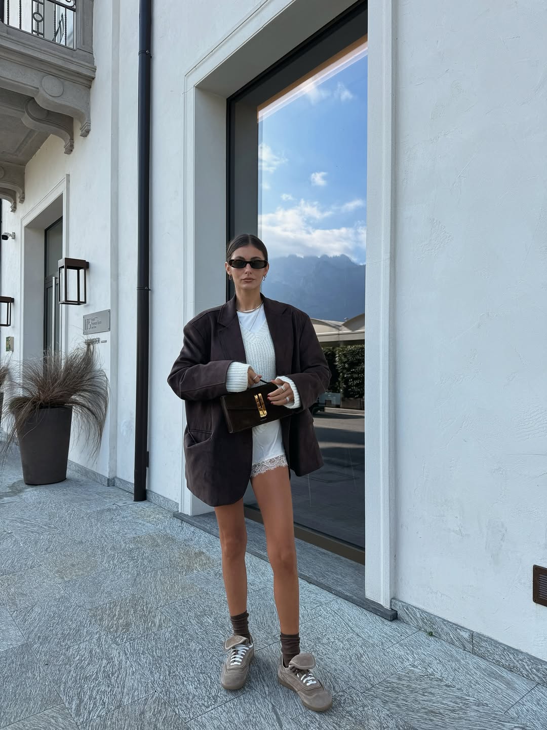 Mulher posando apra foto em cal;ada usando blusa branca, casaco preto e tenis marrom