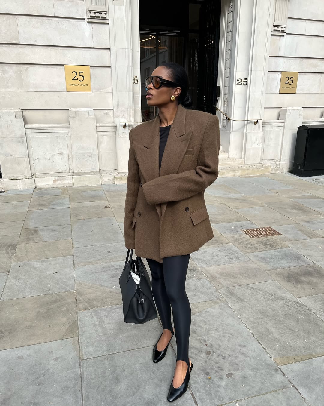 Foto de uma mulher negra usando um look composto por blazer marrom, cal&ccedil;a fus&ocirc;, &oacute;culos de sol, bolsa preta e sapatilha