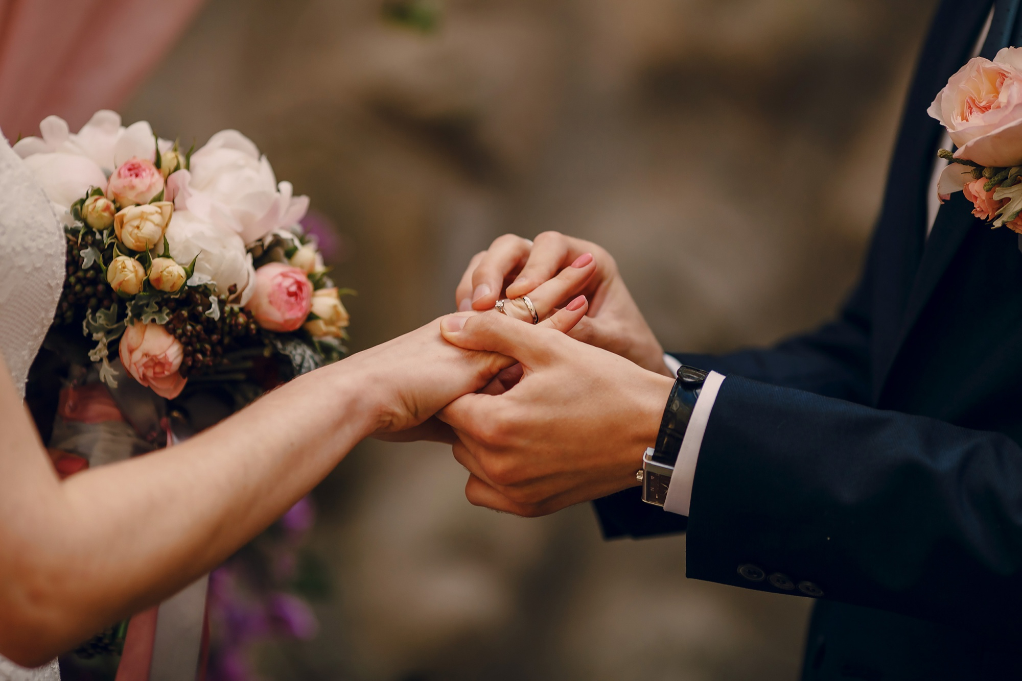 Casal trocando alianças durante cerimônia de casamento, com foco nas mãos e um buquê ao fundo.