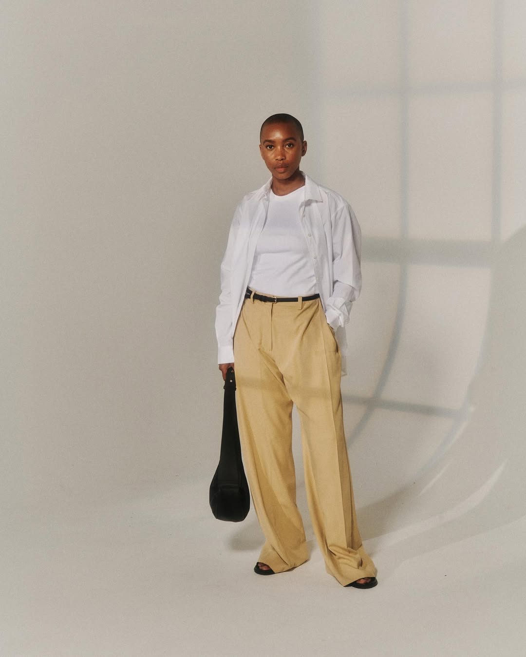 Foto de uma mulher negra usando um look composto por regata branca, cal&ccedil;a de alfaiataria bege, camisa branca e bolsa preta