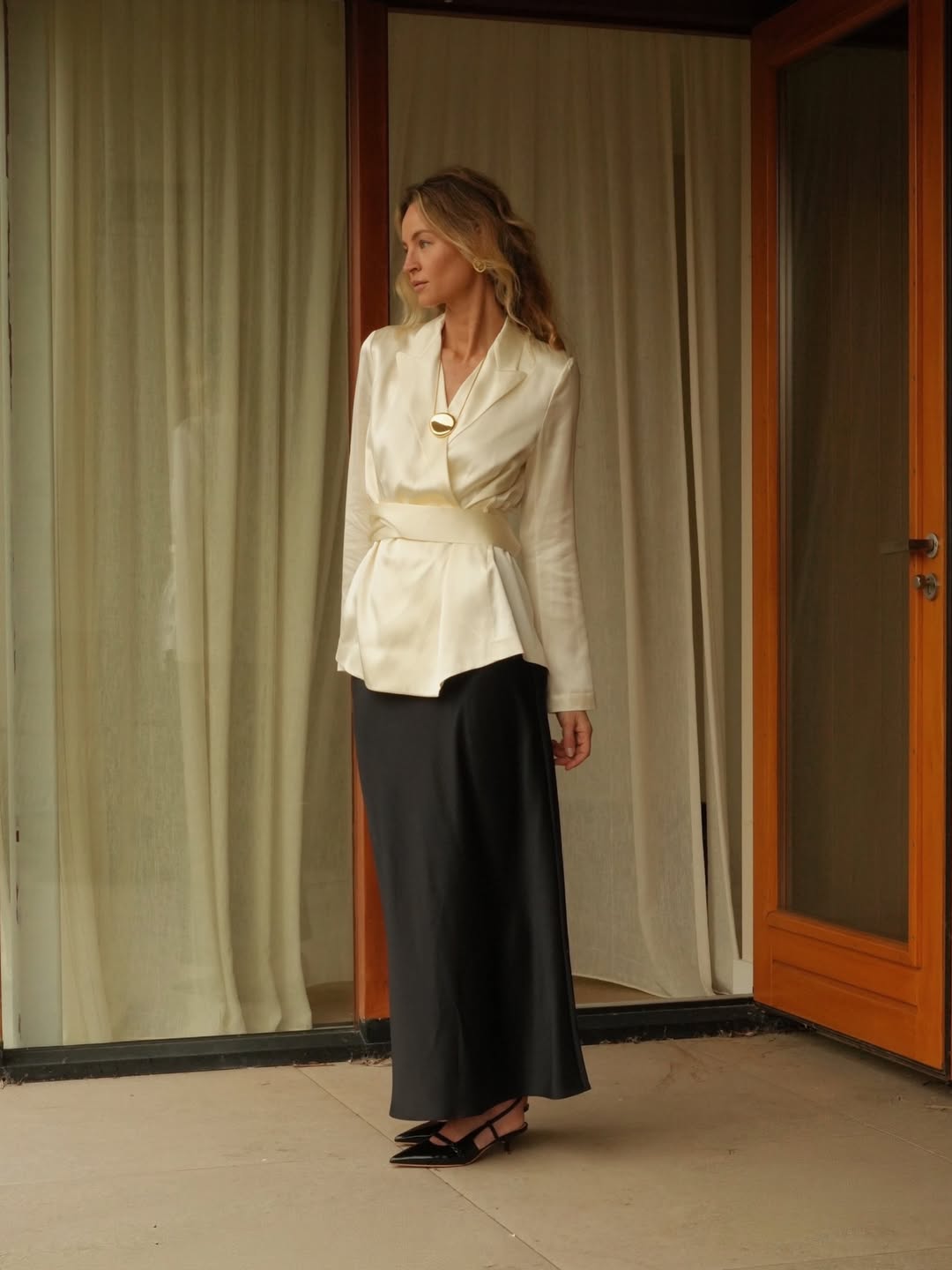 Foto de uma mulher loira usando um look composto por blazer de seda branca, saia longa preta e salto slingback