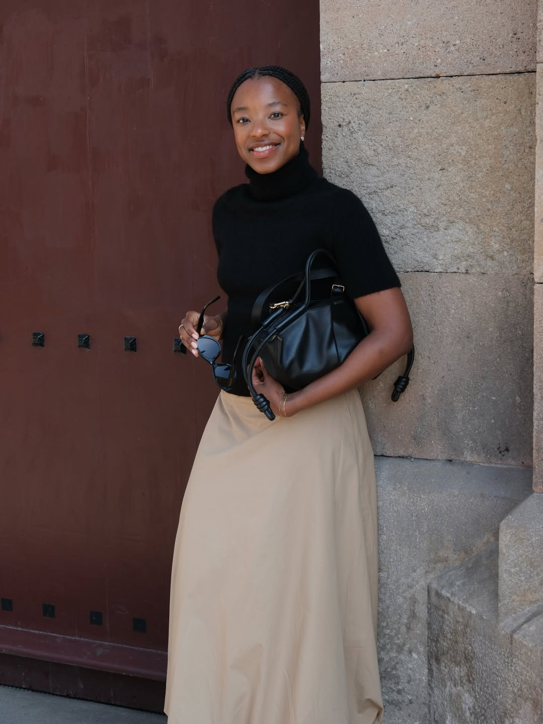 Foto de uma mulher negra usando um look composto por camiseta preta, saia longa de alfaiataria bege e bolsa de couro preta