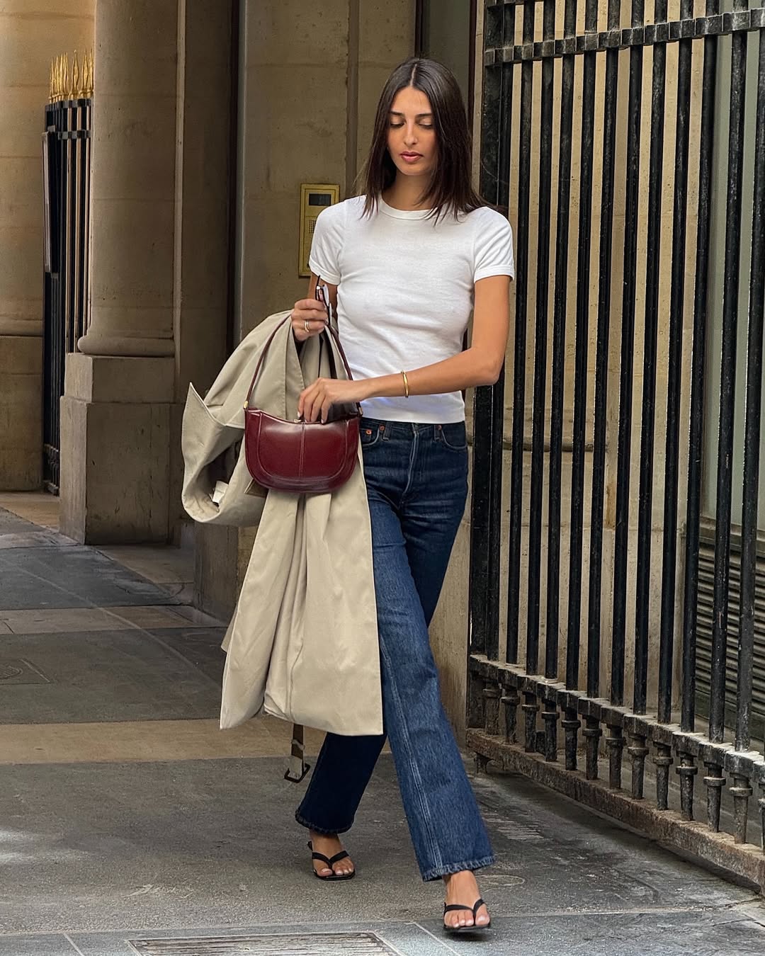 Foto de uma mulher morena usando um look composto por camiseta branca, cal&ccedil;a jeans, bolsa de vinho, trench coat marrom e sand&aacute;lia preta