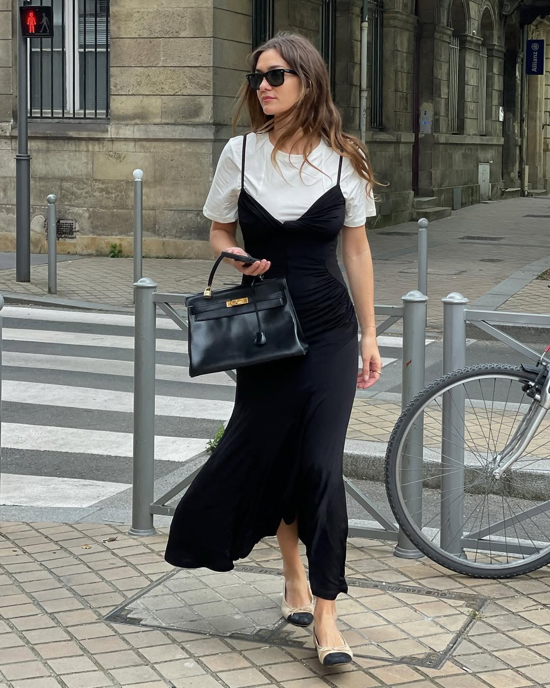 Foto de uma mulher branca usando um look composto por camiseta branca, vestido midi preto, sapatilha e bolsa de couro preta