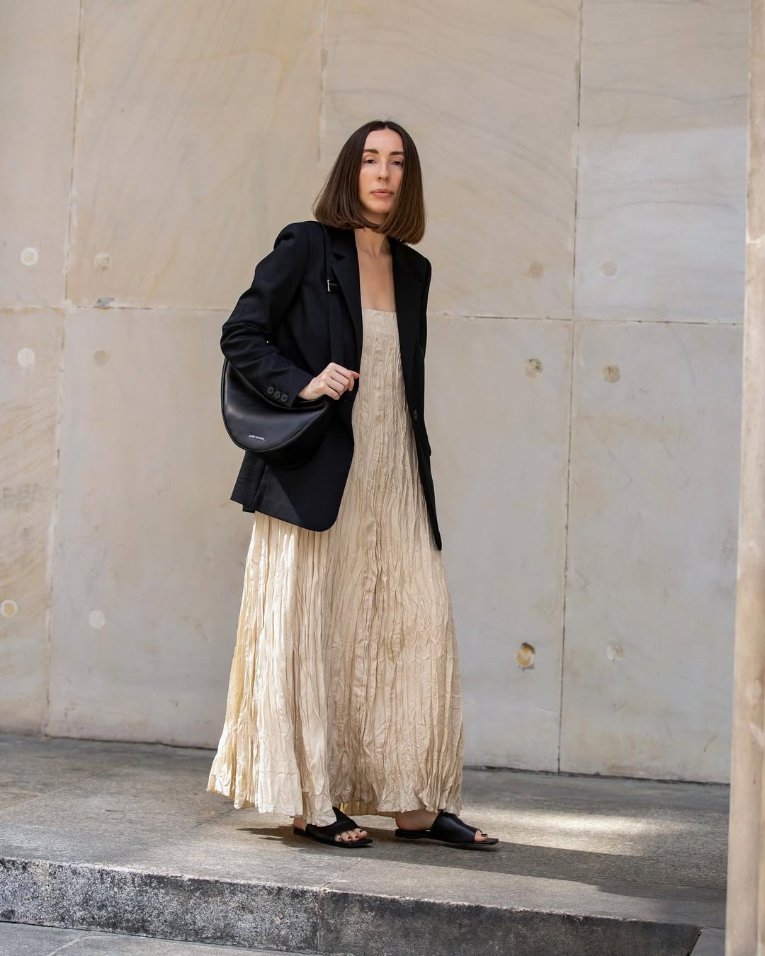 Foto de uma mulher usando um look composto por vestido claro e longo, blazer preto e bolsa de couro