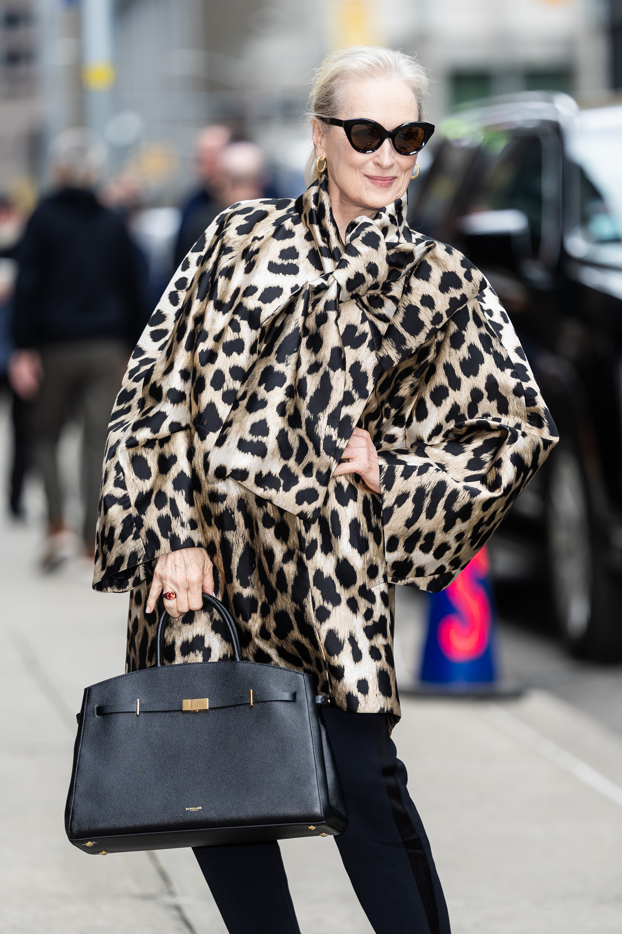 Foto de Meryl Streep. Ela usa um look composto por blusa de oncinha, bolsa preta, cal&ccedil;a preta e slingback preto, al&eacute;m de um &oacute;culos preto em formato de gatinho da Lapima