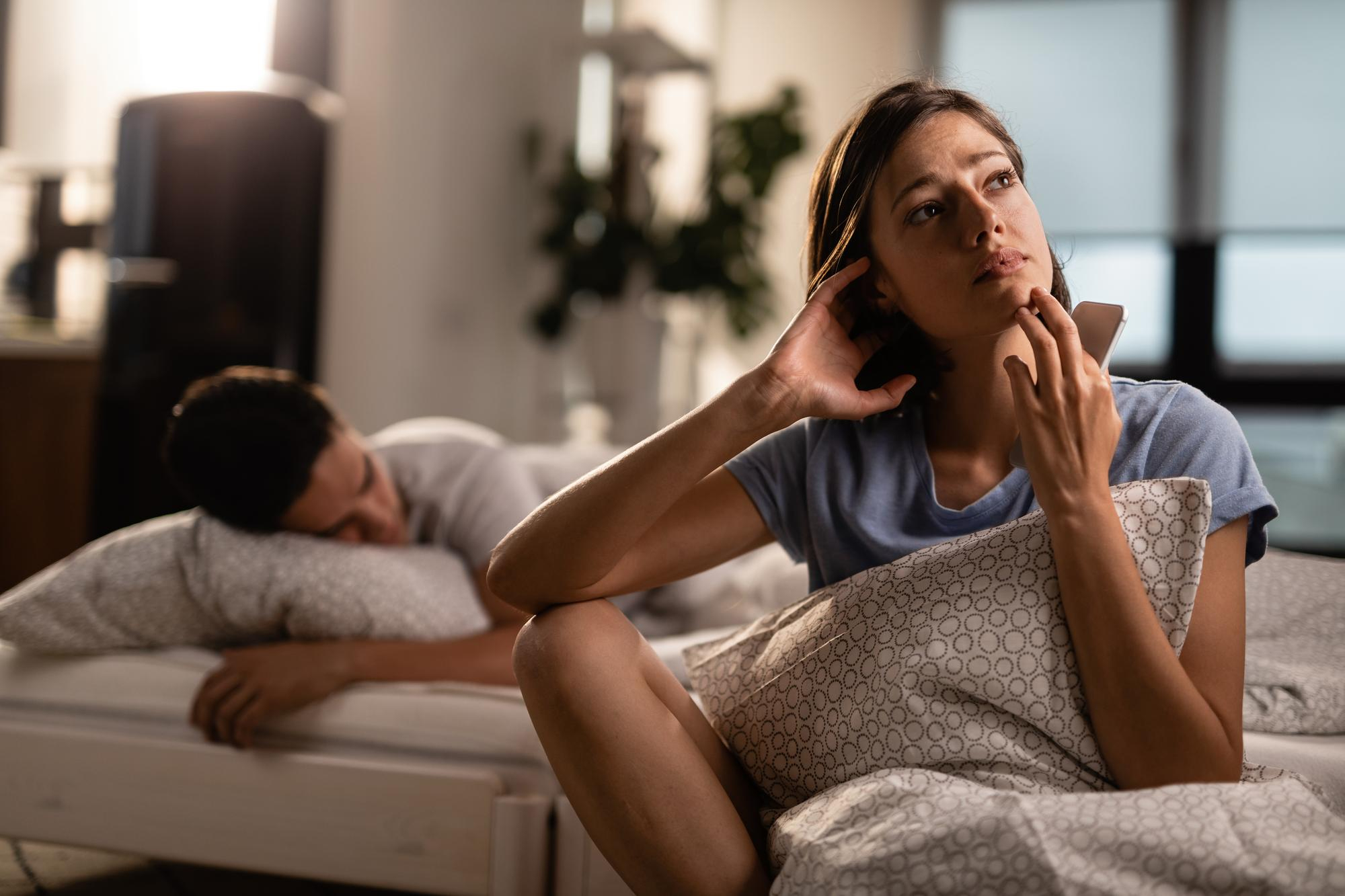 Mulher pensativa segura o celular enquanto parceiro dorme ao fundo, ilustrando falta de conex&atilde;o e esfriamento na rela&ccedil;&atilde;o