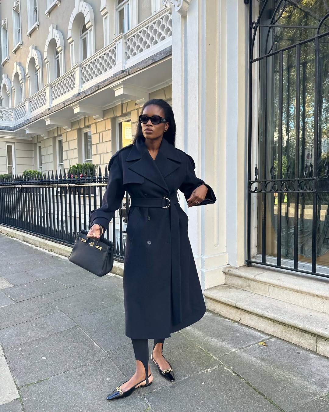 Foto de uma mulher negra usando um look composto por trench coat preto, cal&ccedil;a fus&ocirc;, salto, bolsa e &oacute;culos
