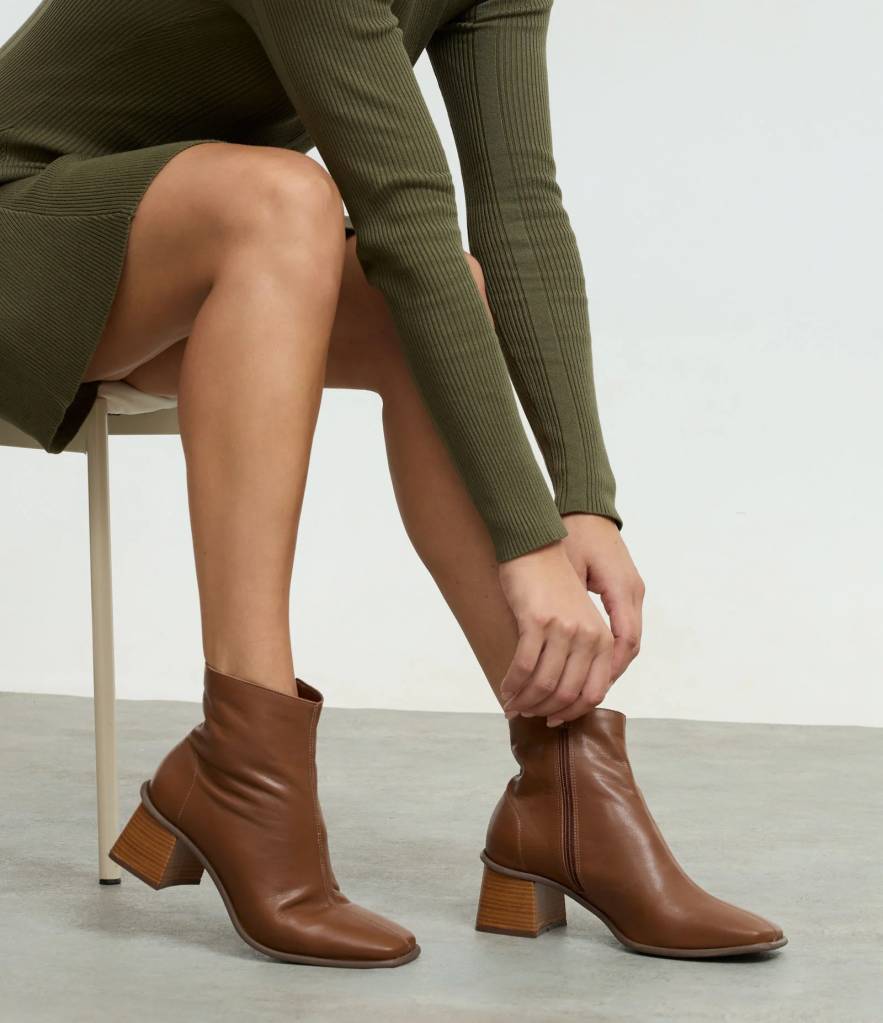 Pessoa sentada em uma cadeira branca, vestindo um vestido verde-oliva canelado, cal&ccedil;ando botas de couro marrom com salto quadrado de madeira, ajustando a bota direita