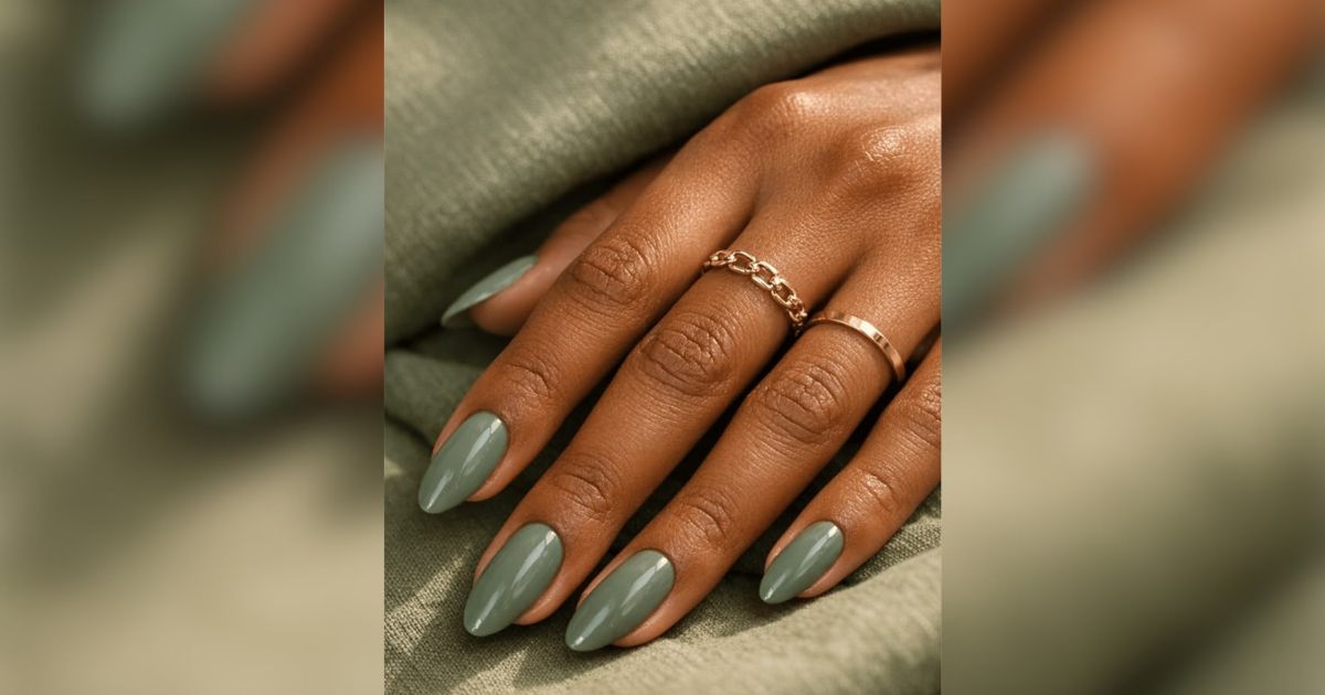 M&atilde;o de pele escura com unhas amendoadas pintadas de verde-oliva, usando dois an&eacute;is dourados, um de corrente e outro liso, sobre tecido verde-musgo