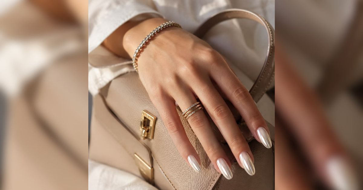 M&atilde;o feminina com unhas longas e peroladas, usando tr&ecirc;s an&eacute;is dourados finos com pedras e uma pulseira de bolinhas prateadas, sobre uma bolsa bege com fecho dourado
