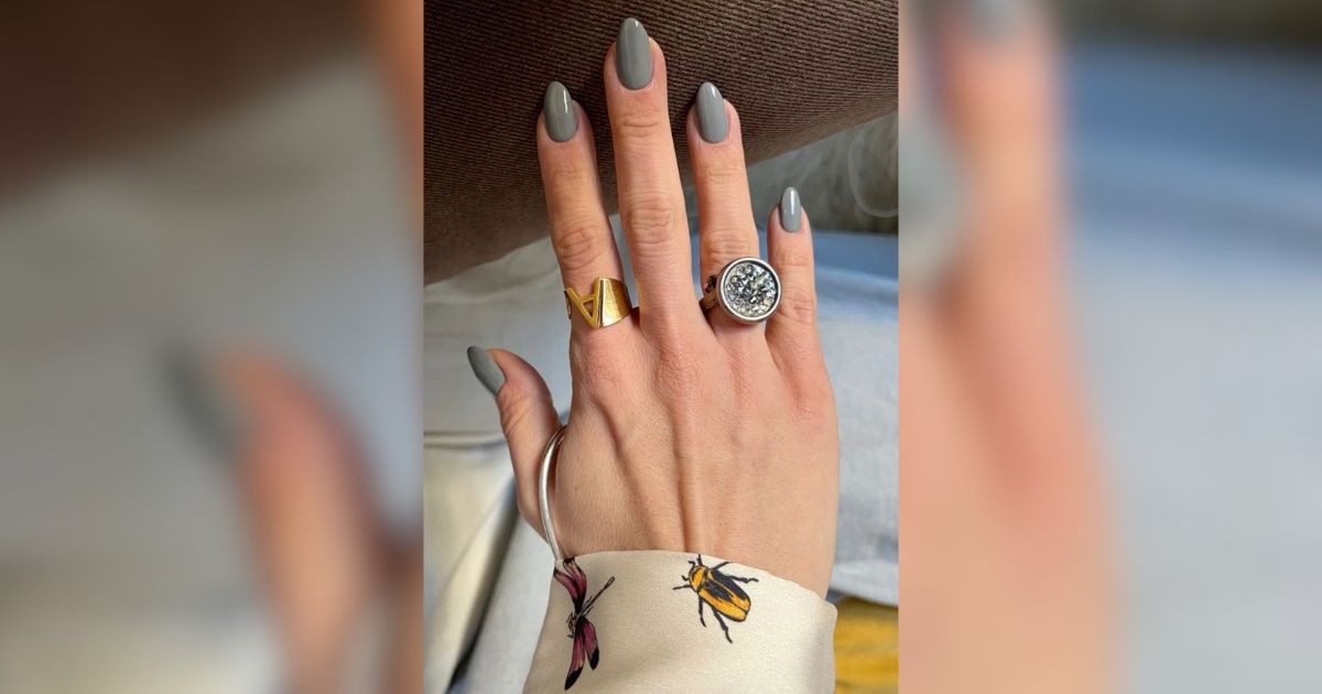M&atilde;o feminina com unhas ovais pintadas de cinza escuro, usando anel dourado com letra 
