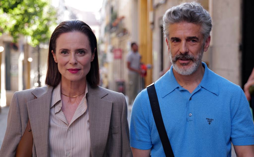 Mulher de cabelo escuro e blazer bege ao lado de homem de cabelo grisalho e barba, vestindo camisa polo azul, em rua com pr&eacute;dios e &aacute;rvores