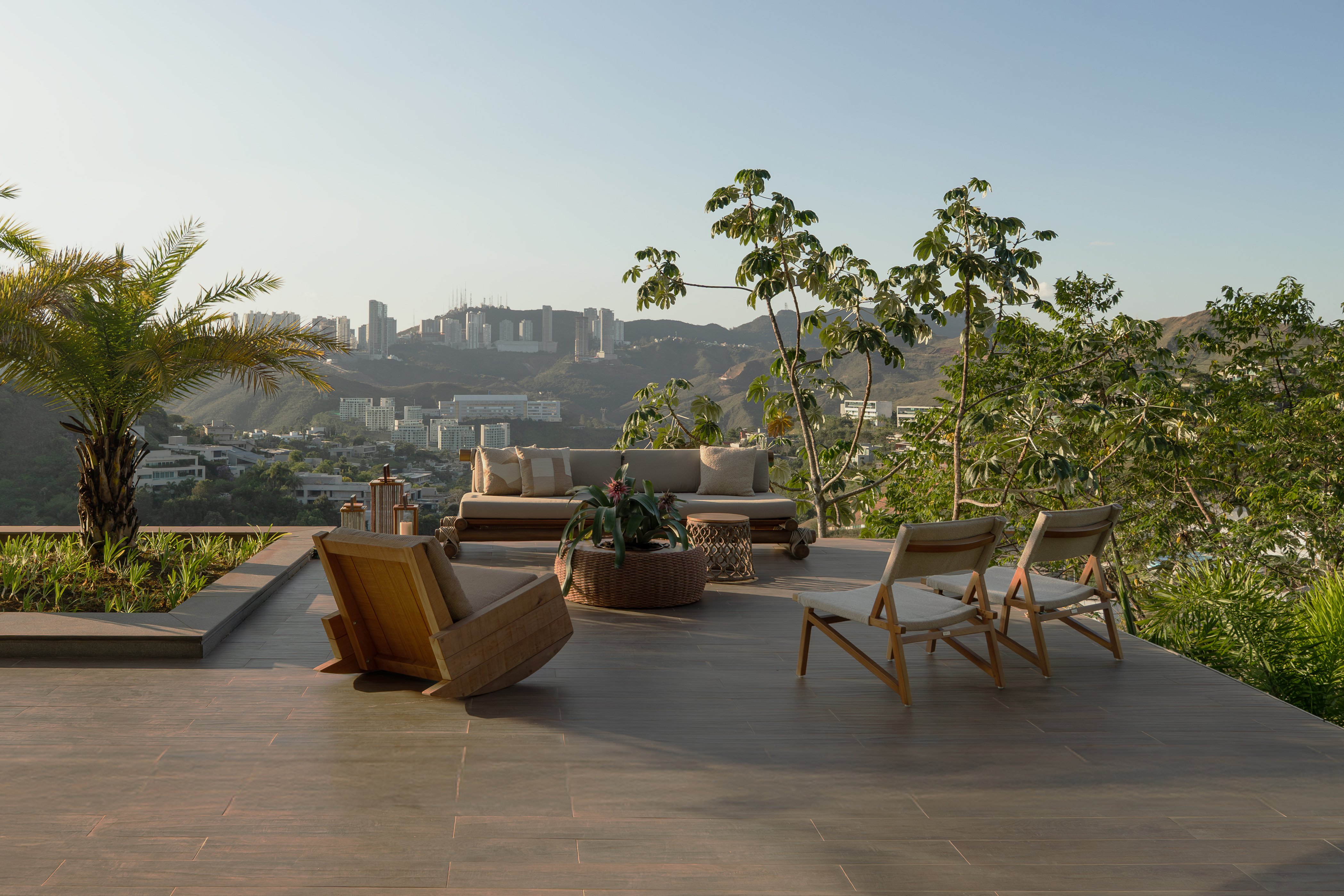 Terra&ccedil;o amplo com piso de madeira, mobili&aacute;rio de &aacute;rea externa e vista para uma cidade em meio a montanhas. H&aacute; um sof&aacute; com almofadas claras, uma poltrona de balan&ccedil;o de madeira e duas cadeiras dobr&aacute;veis com assento de tecido. Plantas e &aacute;rvores verdes emolduram o cen&aacute;rio, com um c&eacute;u claro ao fundo
