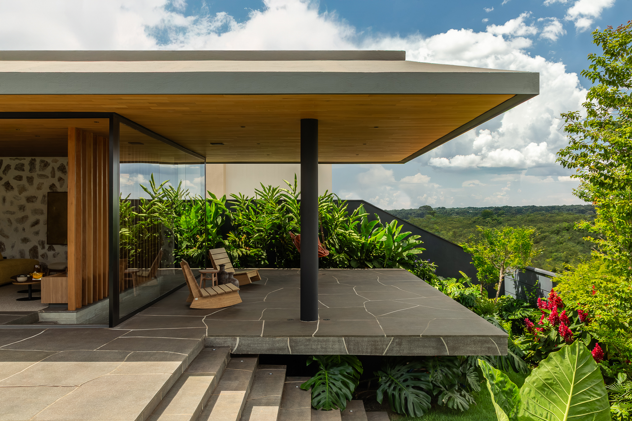 Terra&ccedil;o moderno com piso de pedra escura e duas cadeiras de madeira, cercado por vegeta&ccedil;&atilde;o exuberante e vista para uma floresta sob c&eacute;u azul com nuvens