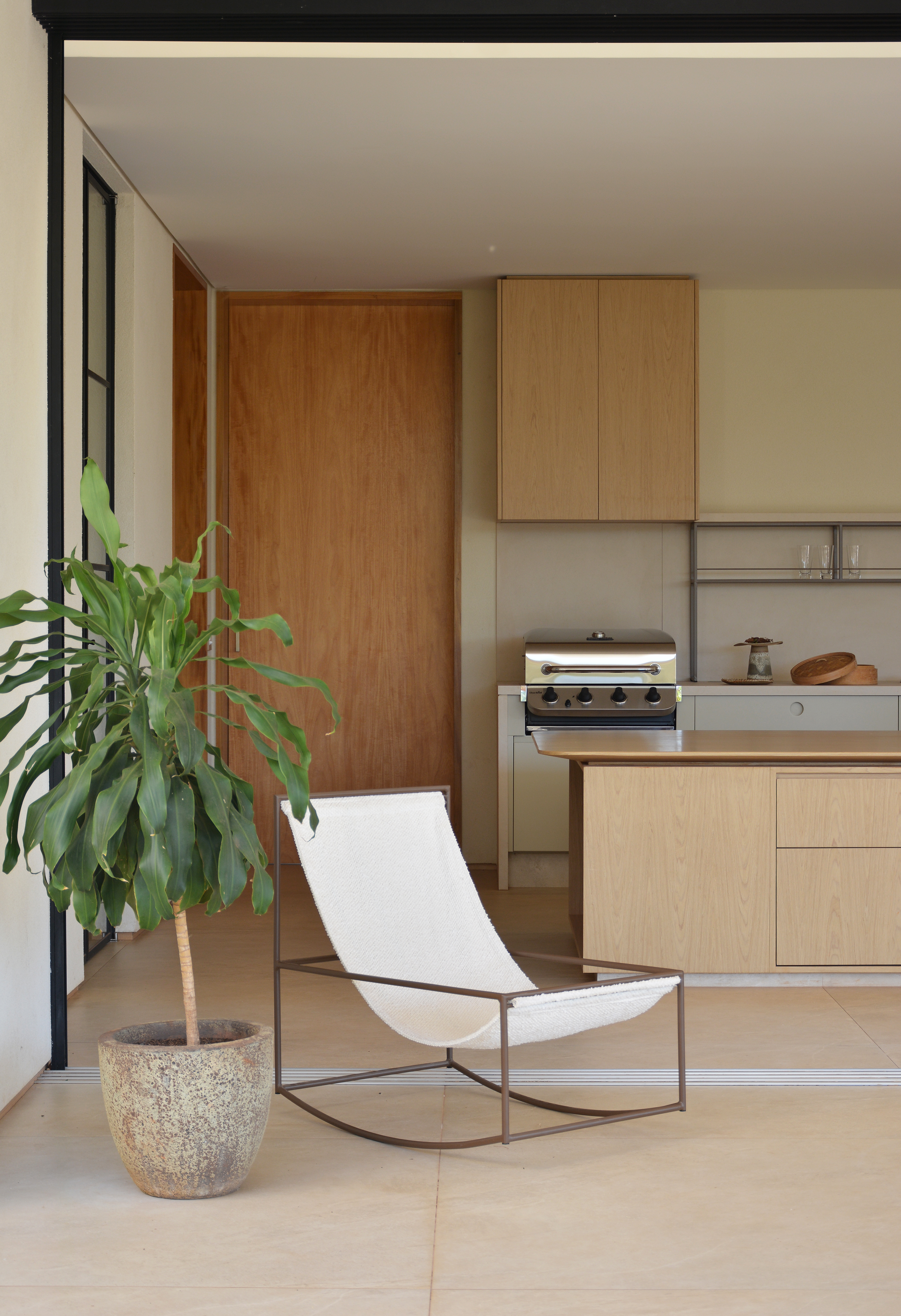 Ambiente interno com cozinha moderna de arm&aacute;rios claros e churrasqueira, uma poltrona de balan&ccedil;o branca e um vaso de planta verde no primeiro plano