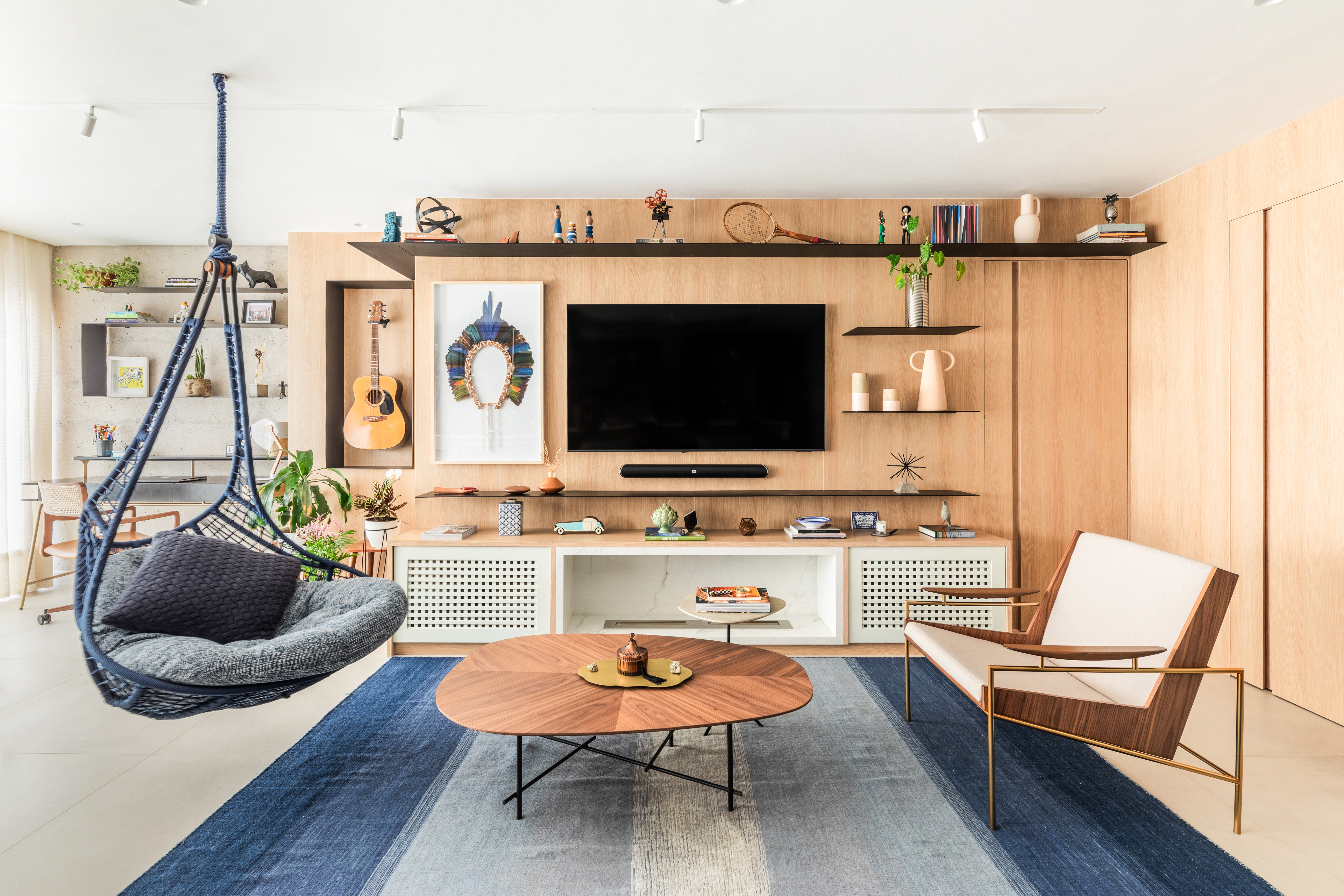 Sala de estar moderna com parede de madeira clara, TV preta centralizada, prateleiras com objetos decorativos e um viol&atilde;o. &Agrave; esquerda, uma cadeira suspensa azul com almofadas cinzas. No centro, um tapete azul e branco, e uma mesa de centro oval de madeira. &Agrave; direita, uma poltrona de madeira e estofado branco. O ambiente &eacute; iluminado e acolhedor