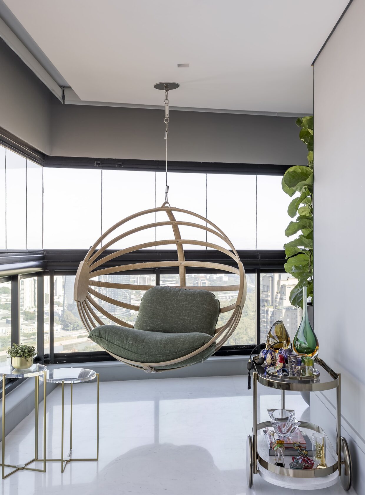 Varanda gourmet com poltrona suspensa de madeira clara e almofada verde, vista panor&acirc;mica da cidade, duas mesas laterais redondas e um carrinho de bar com garrafas e copos
